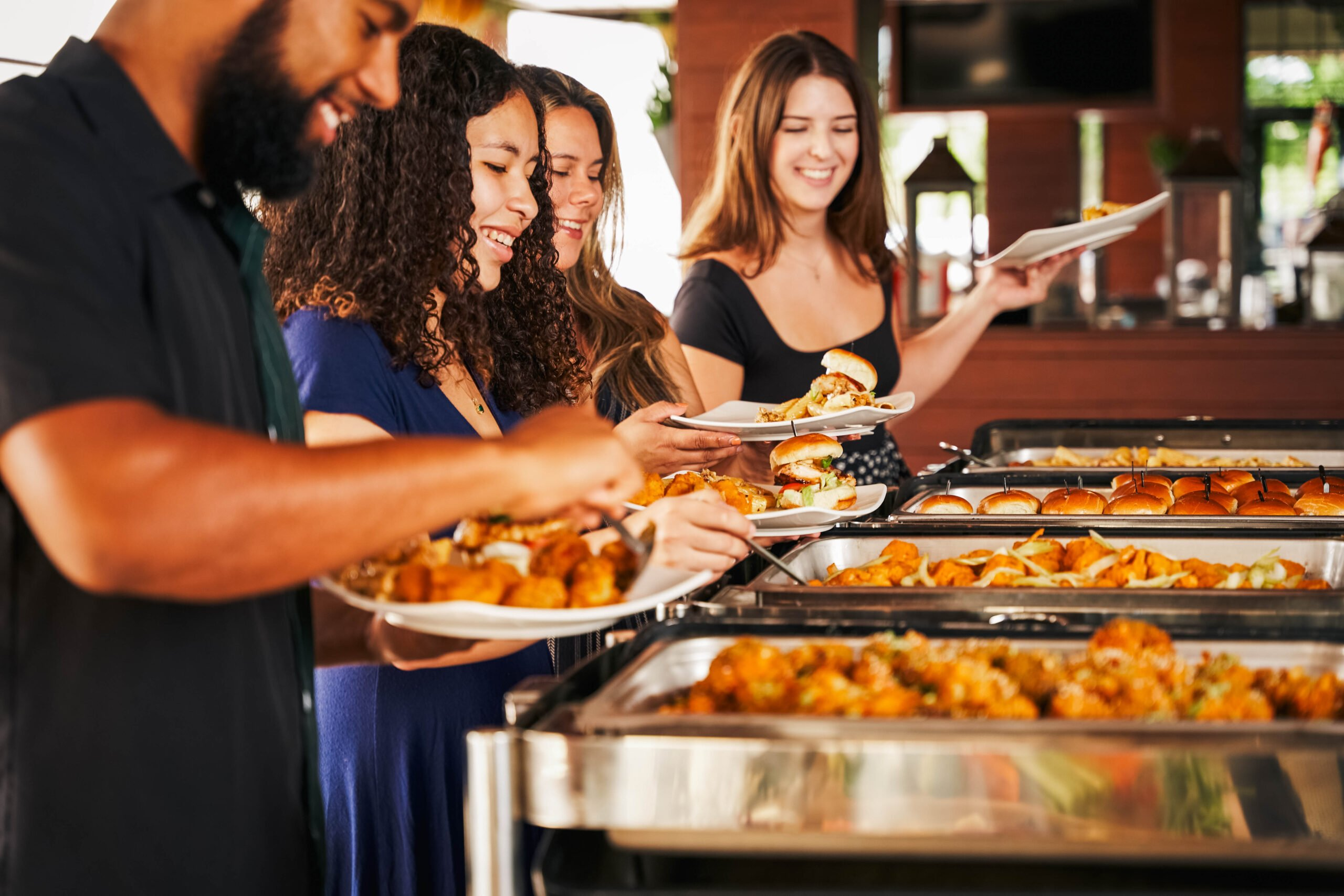 Four people serve themselves food from a buffet line, holding plates and smiling in a casual indoor setting at an American Tavern Bar & Restaurant.