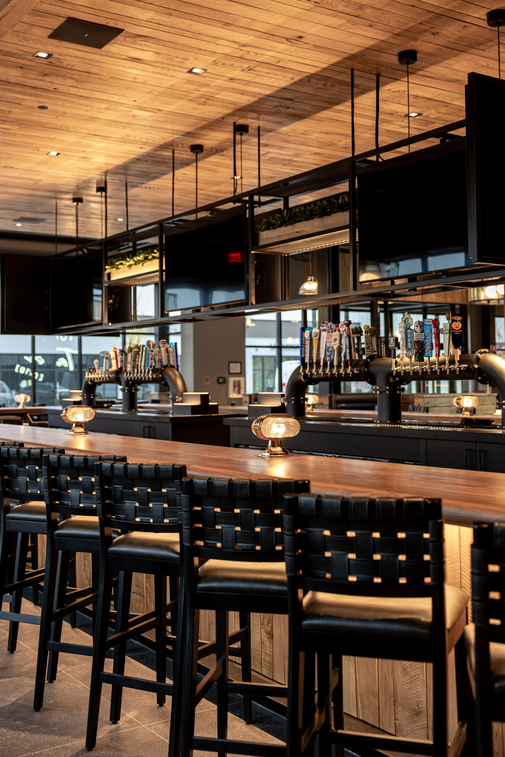 A modern Bar & Restaurant interior with high wooden stools, a long counter, overhead shelving, multiple beer taps, and warm ambient lighting inspired by an American Tavern.