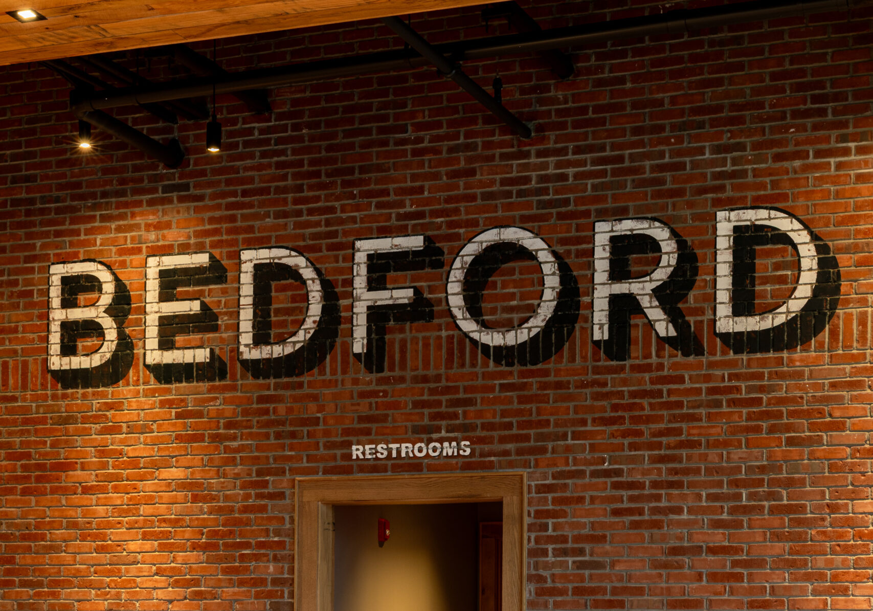 A row of high-backed bar stools lines a wooden bar in front of a brick wall, evoking the classic charm of an American Tavern. Above a doorway, “BEDFORD” and a “Restrooms” sign add character to this inviting Bar & Restaurant setting.