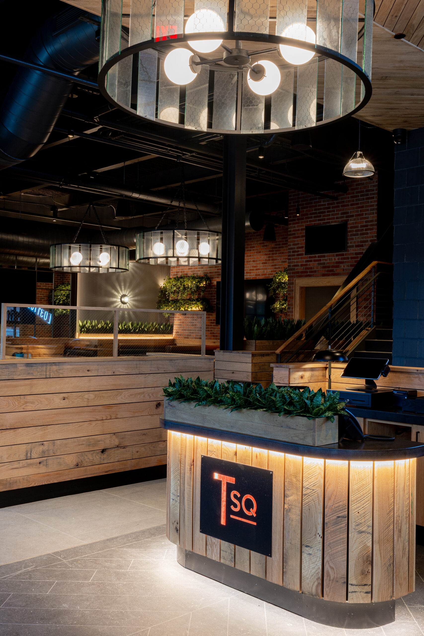 Modern American Tavern reception area with a circular wooden desk featuring an illuminated “TSQ” sign, hanging light fixtures, wood paneling, and lush plants—setting the tone for this inviting bar & restaurant.