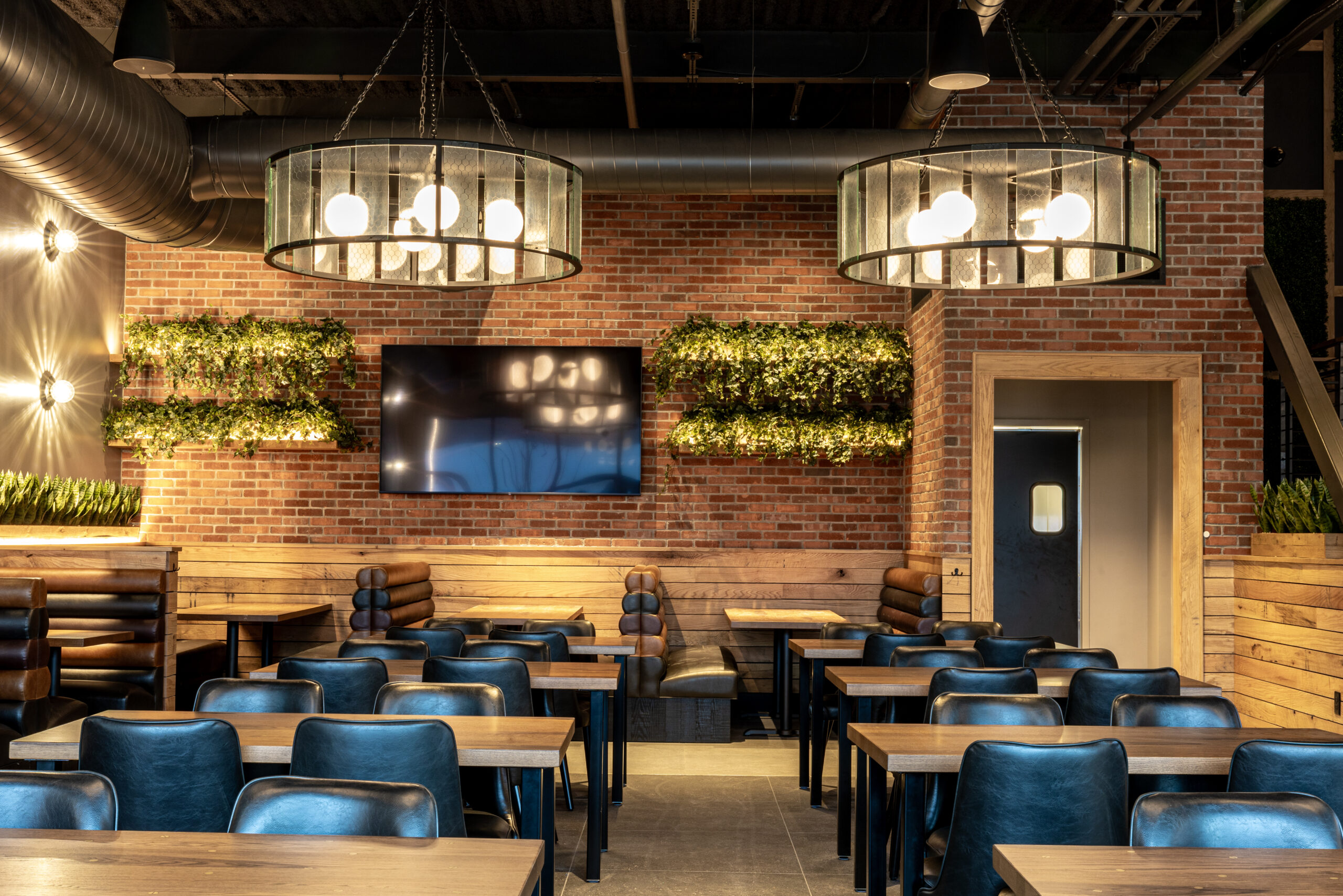 A modern American Tavern dining area with wooden tables, blue chairs, booth seating, a large TV on a brick wall, green plant decor, and two round chandeliers.