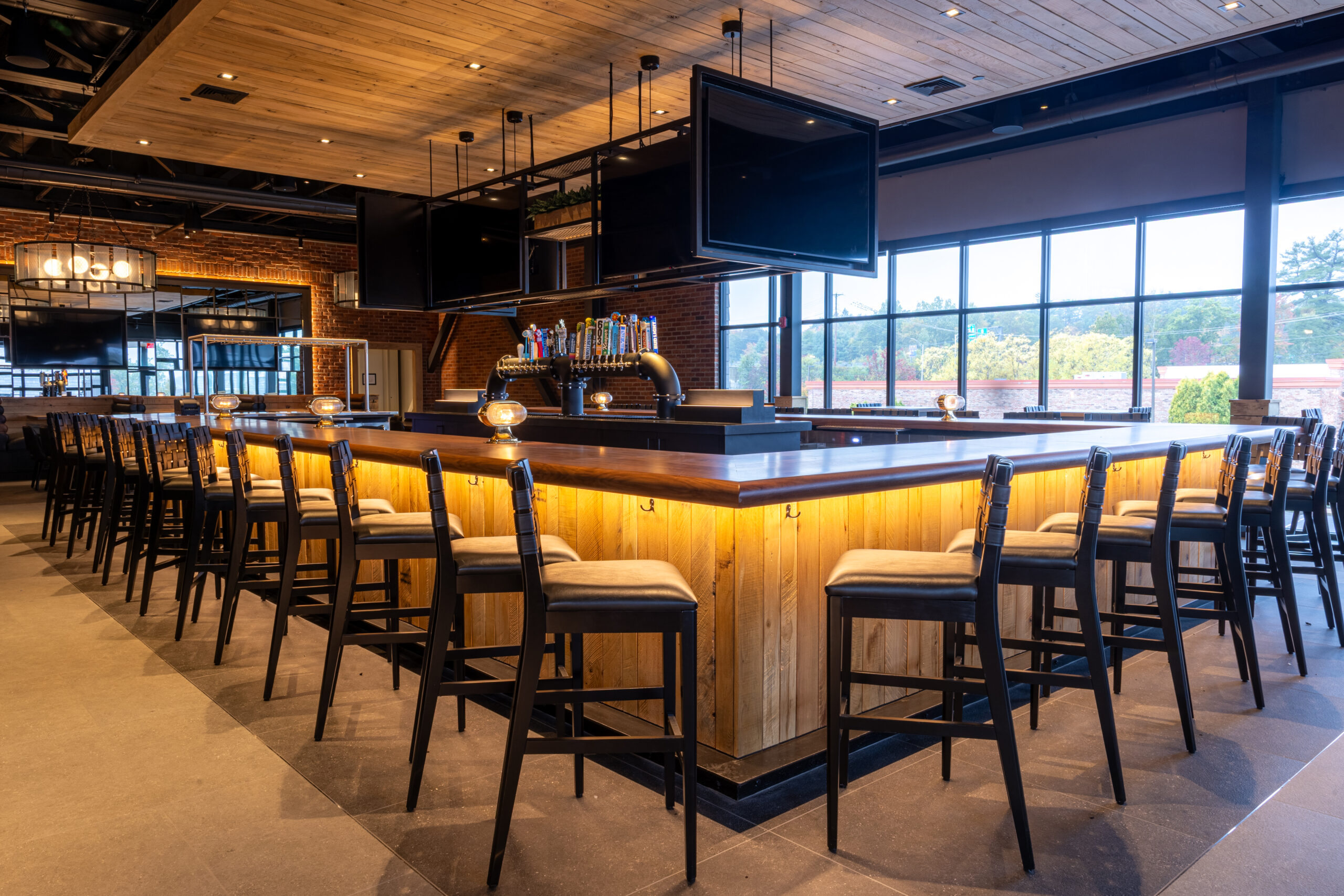 A modern American Tavern, this spacious bar & restaurant features wooden accents, high chairs around the counter, beer taps, and multiple ceiling-mounted TVs, all set in a bright room with large windows.
