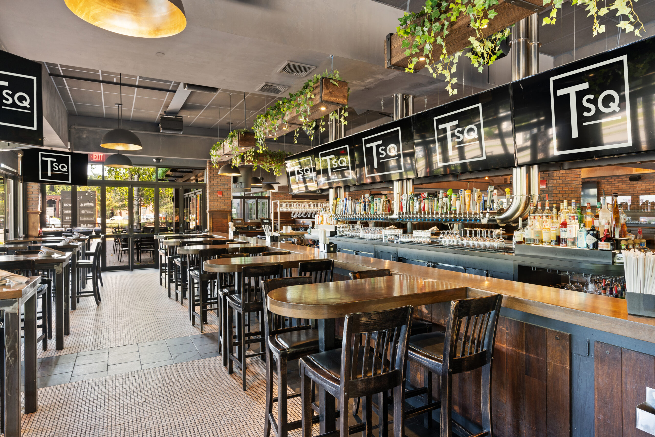 Modern Bar & Restaurant interior with wooden tables, high chairs, a long bar with taps, hanging plants, and large TSq logo signs—this American Tavern welcomes natural light from expansive windows.