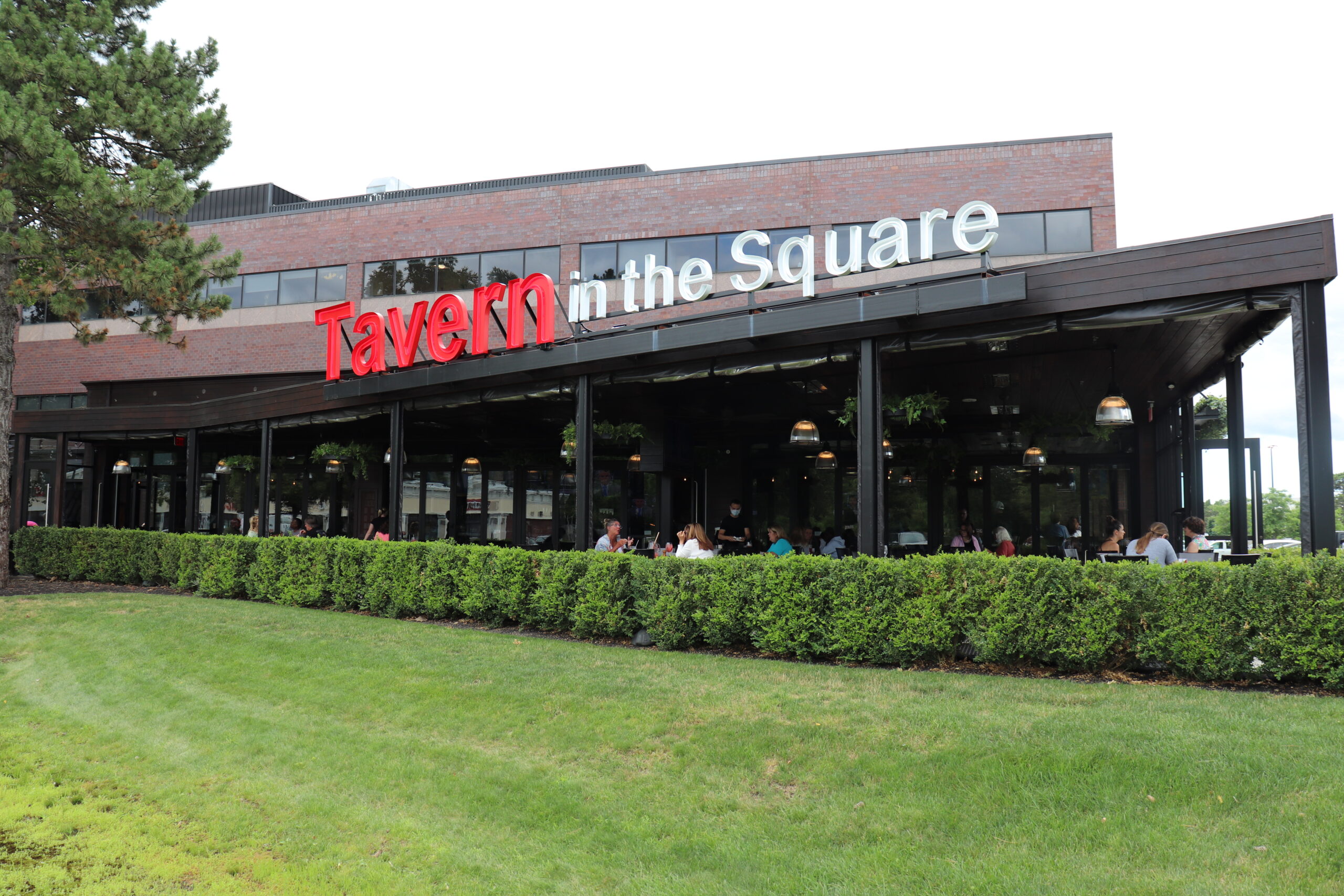 People sit at outdoor tables under a covered patio at Tavern in the Square, an American Tavern & Bar Restaurant with a brick exterior and large windows.