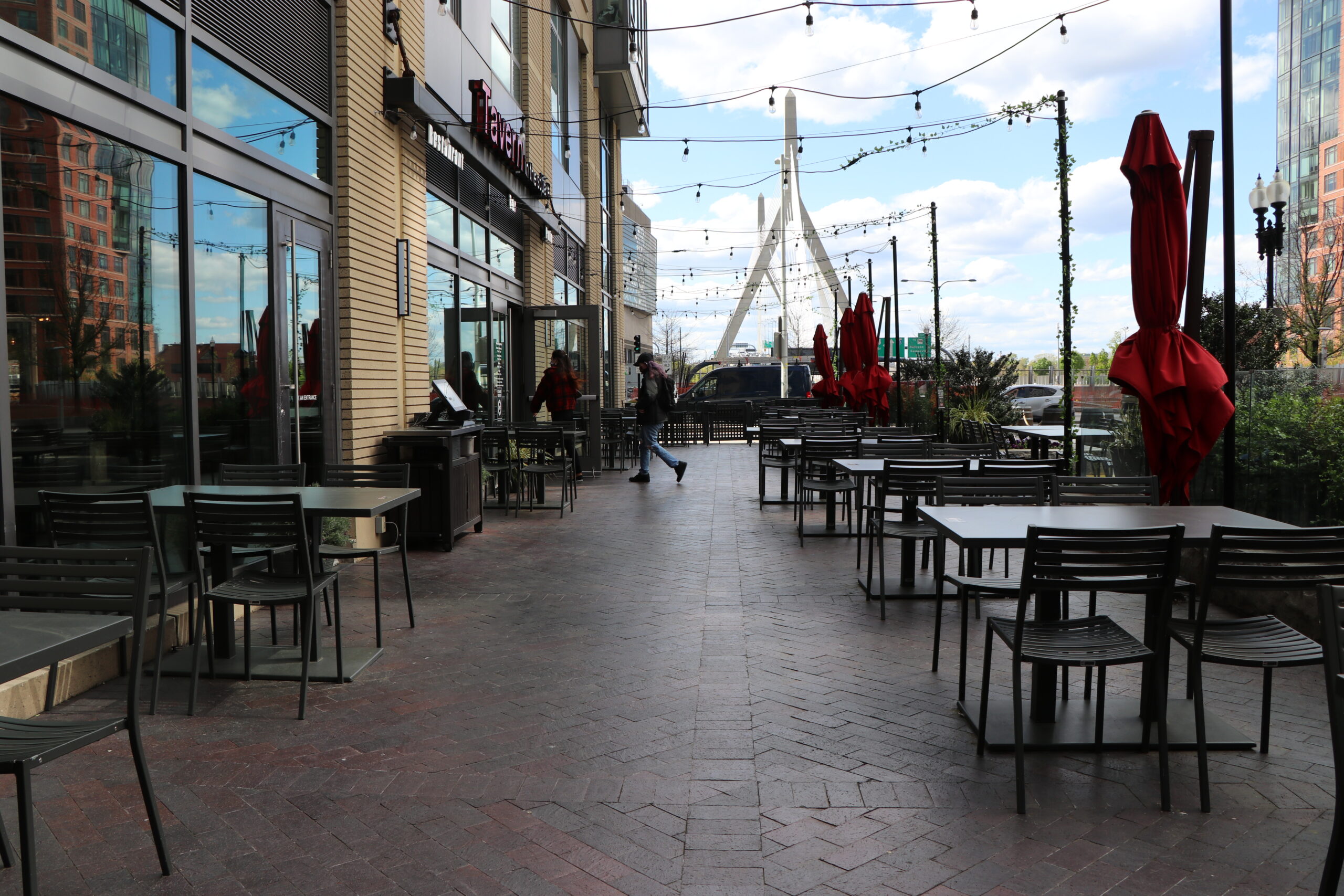 Outdoor Bar & Restaurant patio with empty tables and chairs, closed red umbrellas, and string lights; a person walks near the entrance of this inviting American Tavern on a partly cloudy day.