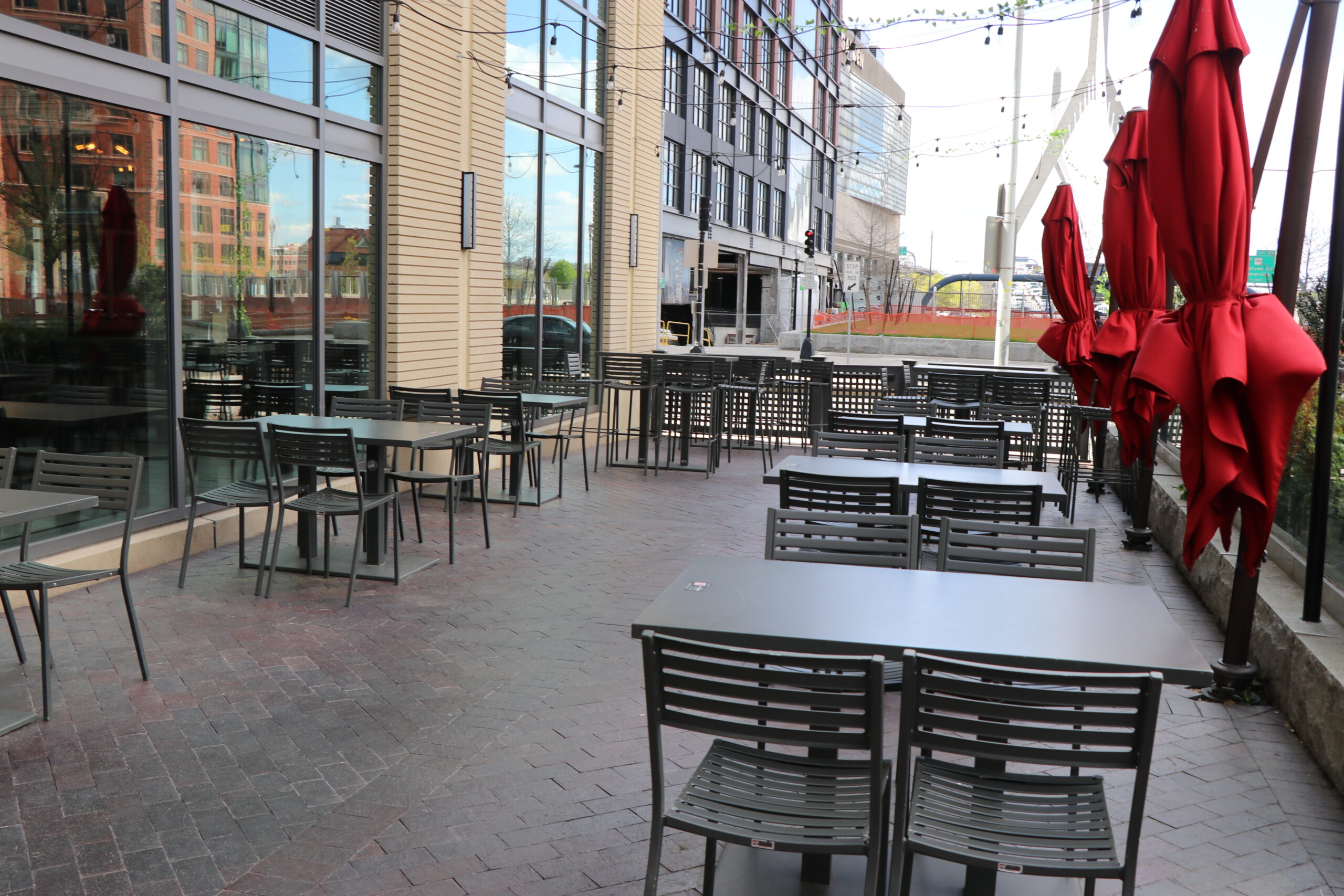 Outdoor patio area at this American Tavern Bar & Restaurant features empty metal tables and chairs, several closed red umbrellas, and a building with large windows in the background.