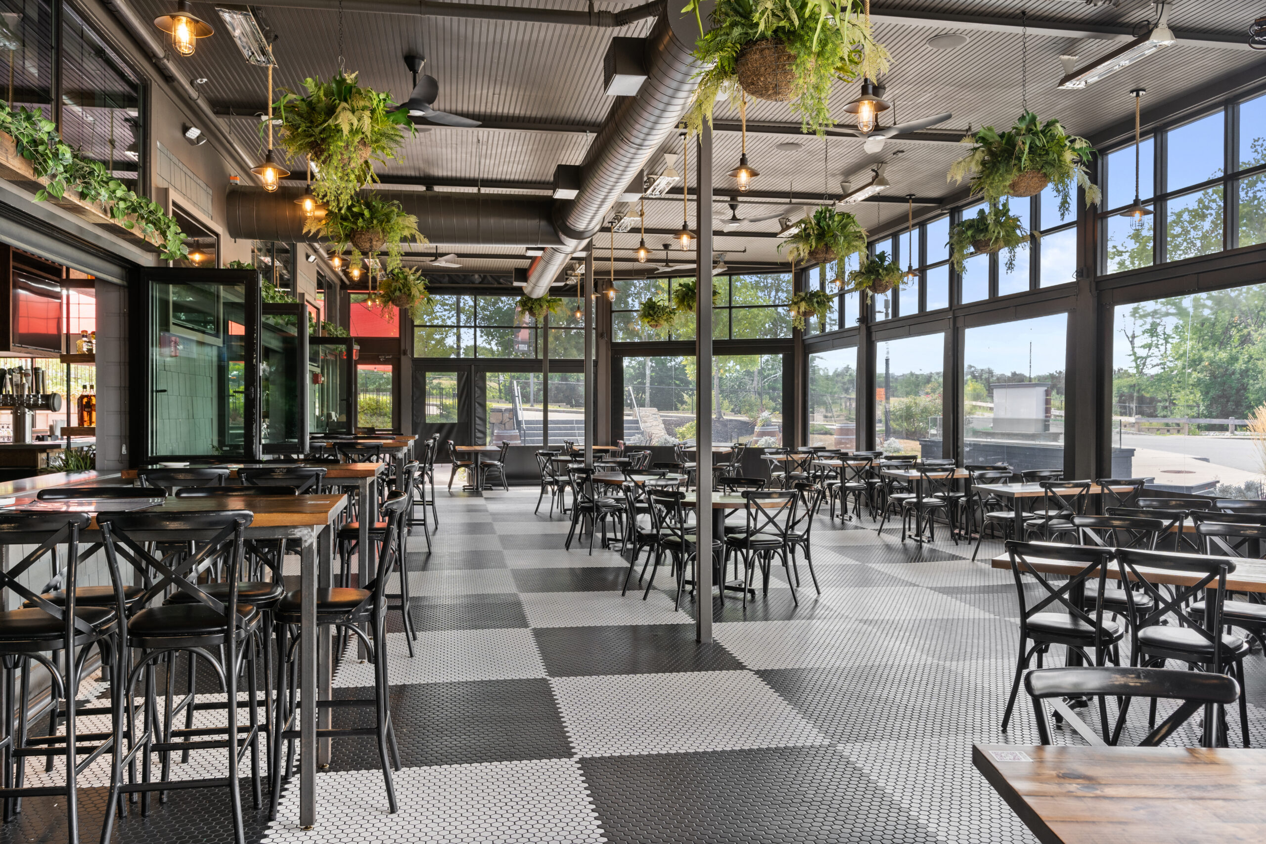 Spacious American Tavern with black metal chairs, wooden tables, large windows, hanging plants, checkered floor, and abundant natural light.