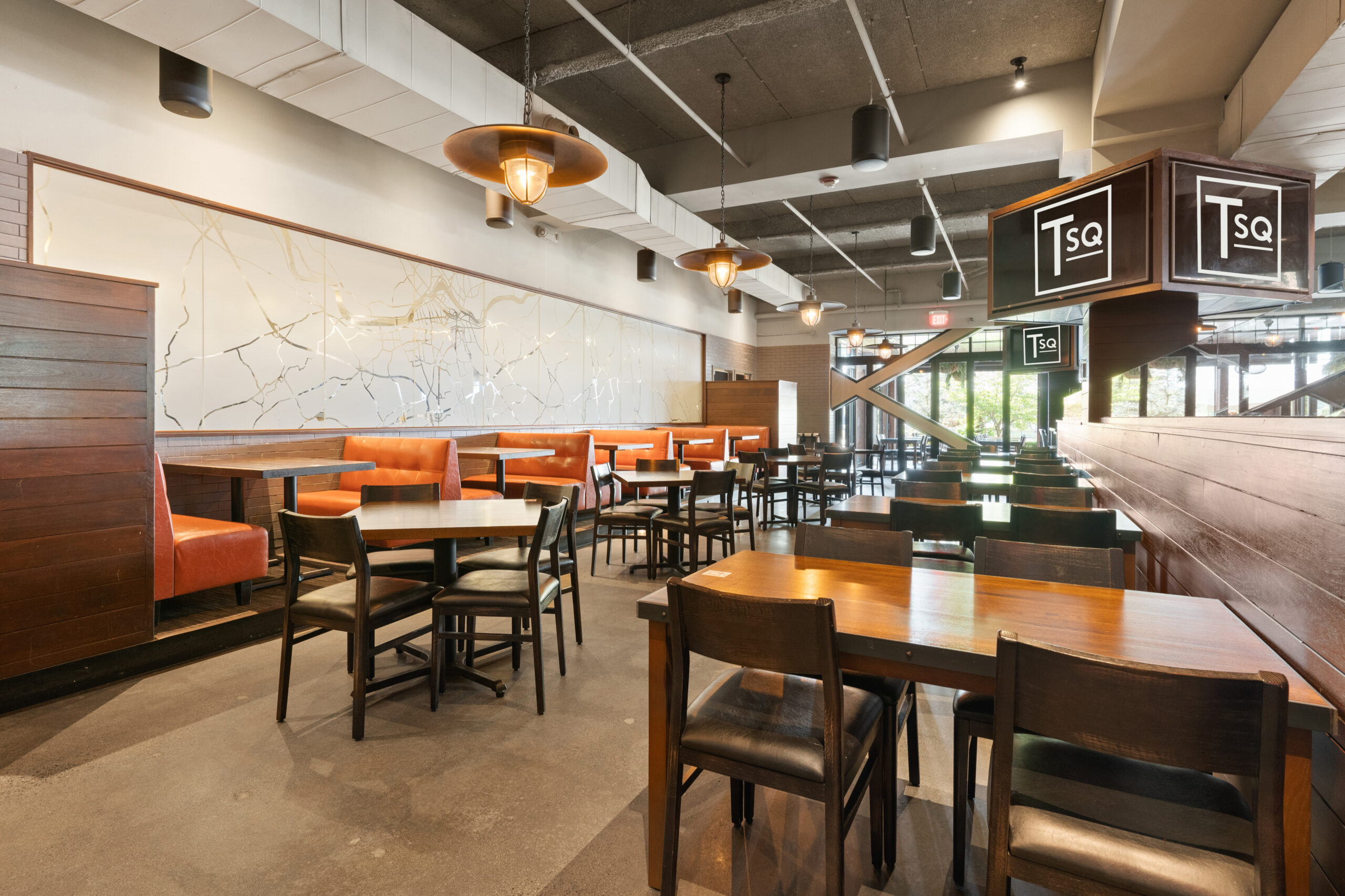 Modern American Tavern interior featuring wooden tables and chairs, orange upholstered booth seating, large wall art, and natural light streaming through tall windows in this inviting bar & restaurant.