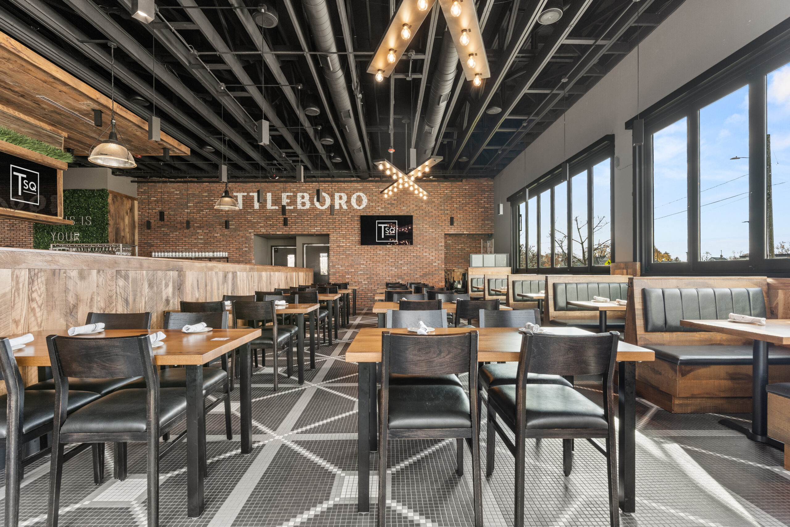 Modern Bar & Restaurant interior with wooden tables, black chairs, booth seating, geometric tile floor, exposed ceiling, and large windows allowing natural light—perfect for an inviting American Tavern atmosphere.