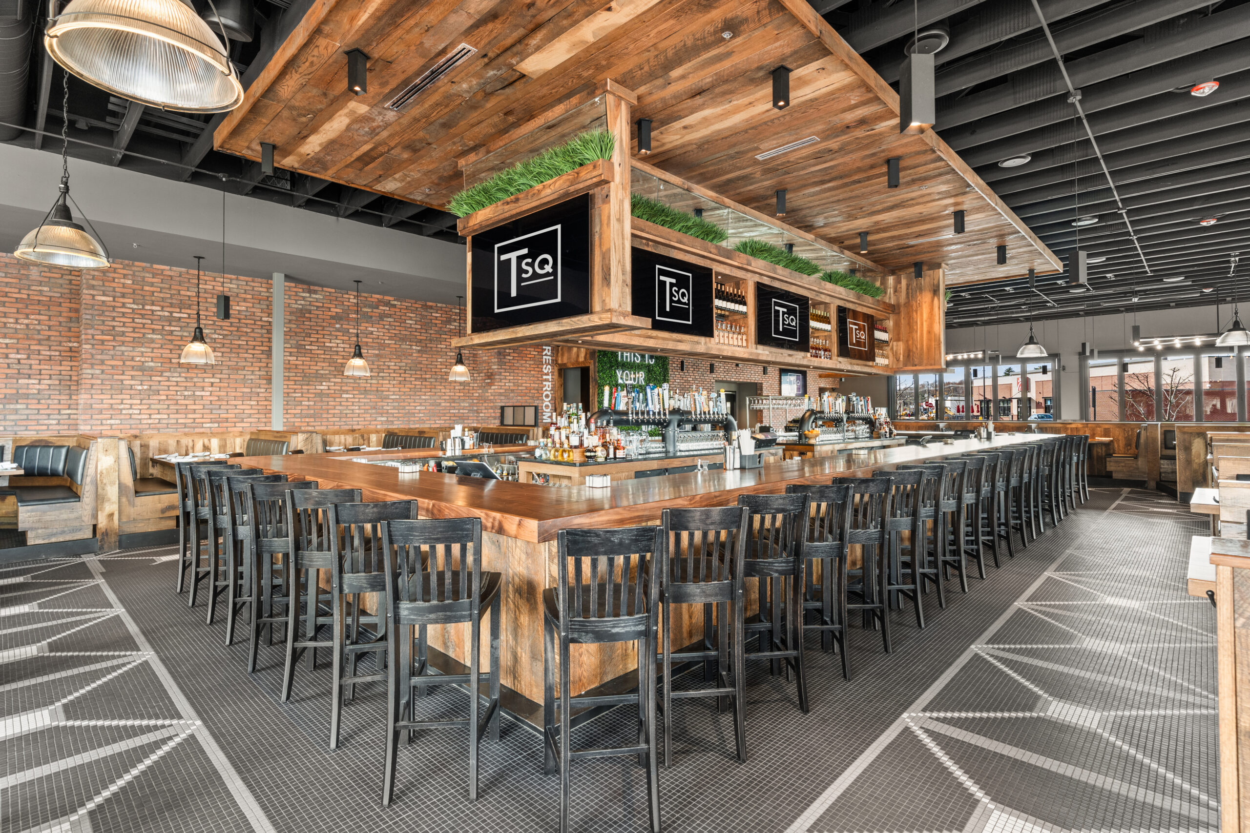 A modern American Tavern featuring a large wooden U-shaped counter, high black chairs, multiple taps, overhead signs, exposed brick walls, and industrial lighting—perfect for a classic Bar & Restaurant experience.