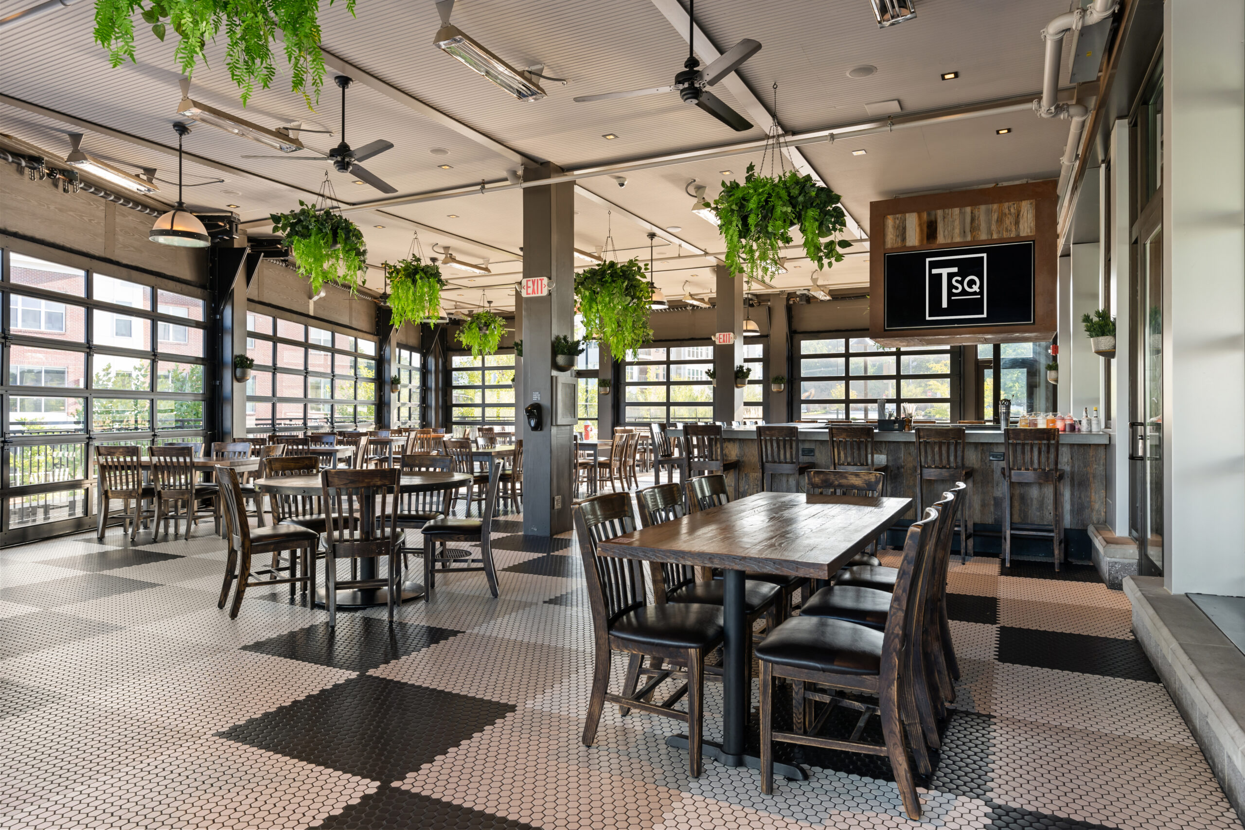 Spacious American Tavern interior with wooden tables and chairs, potted plants hanging from the ceiling, large windows, and a tiled floor makes this Bar & Restaurant warm and inviting.