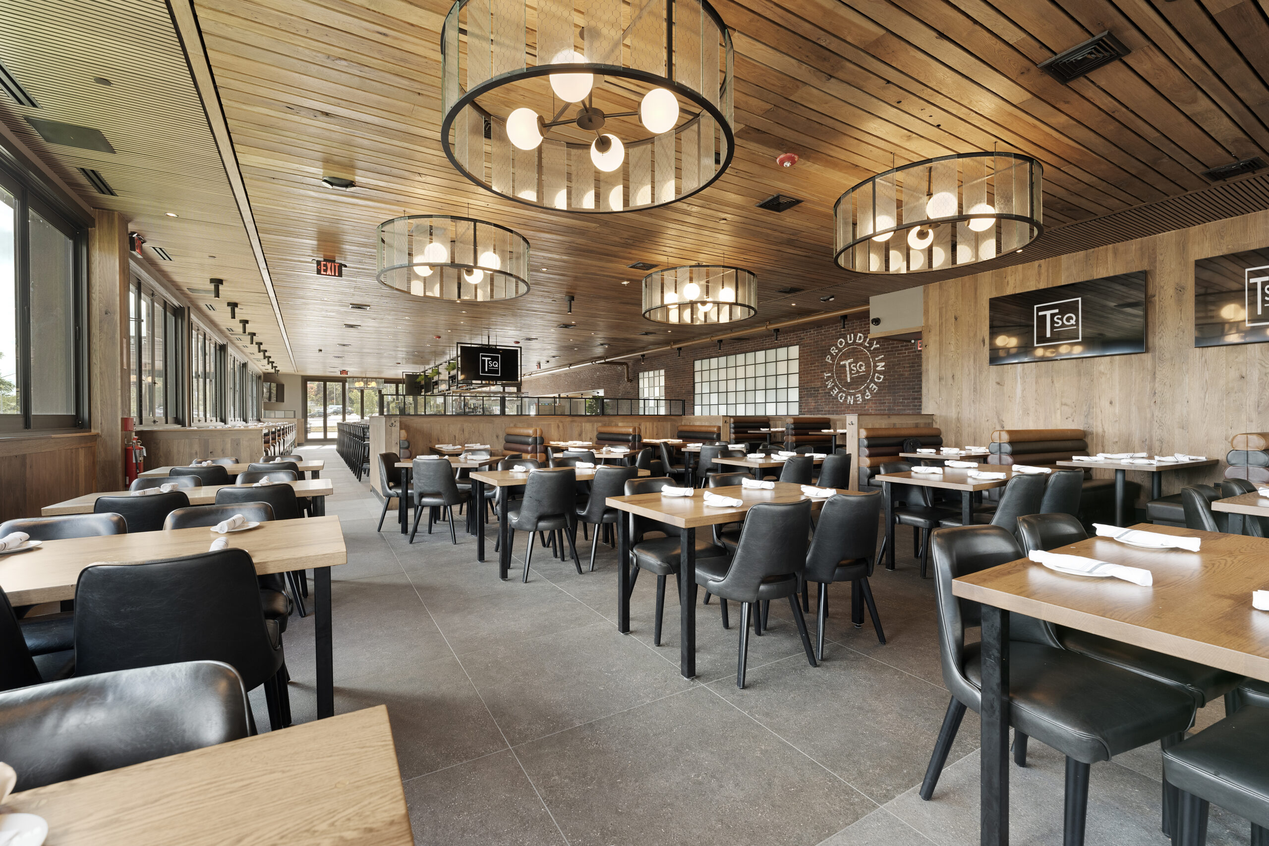 Modern American Tavern interior with wooden ceiling, pendant lights, black chairs, and wooden tables set for dining. Large windows line one side, and décor includes wall logos and TV screens in this inviting bar & restaurant.