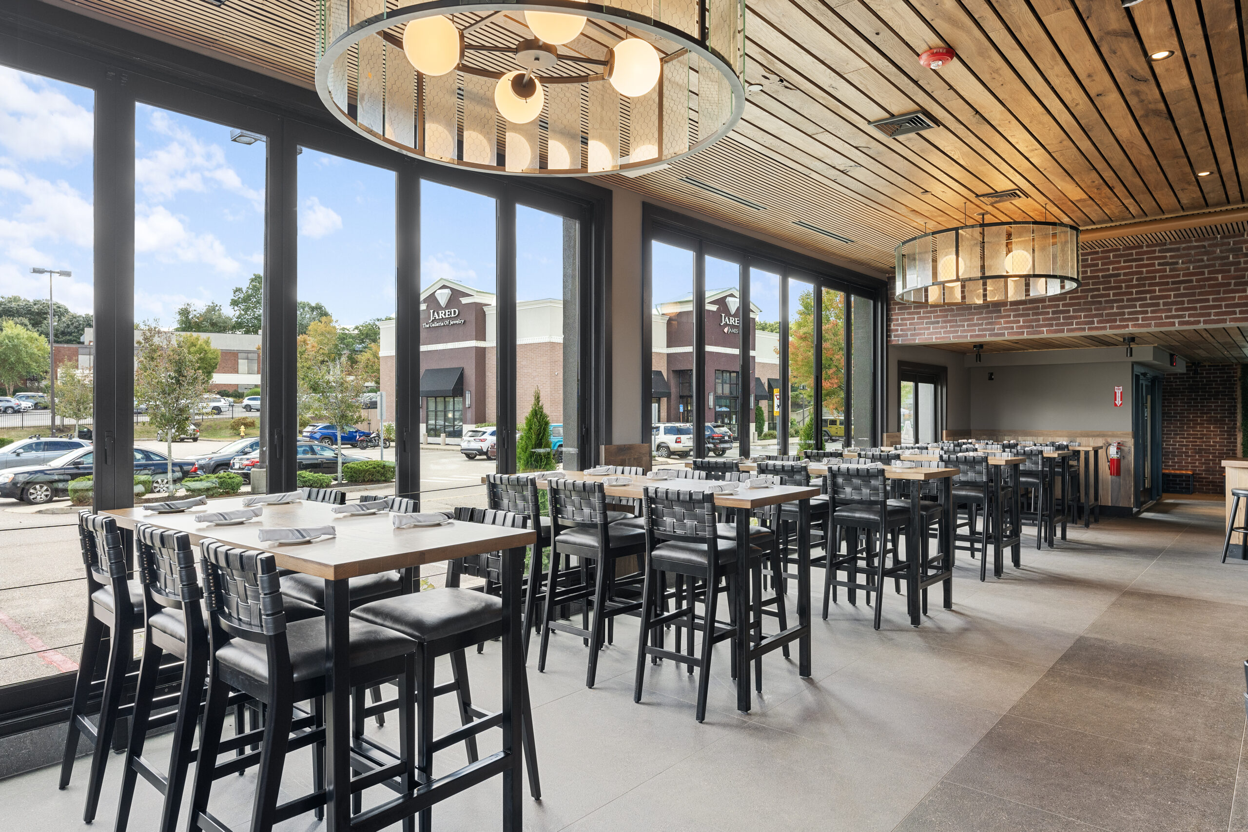 Modern Bar & Restaurant interior with high tables and chairs, large windows overlooking a shopping center, and ceiling lights. Daylight streams in through the windows, creating a welcoming American Tavern atmosphere.
