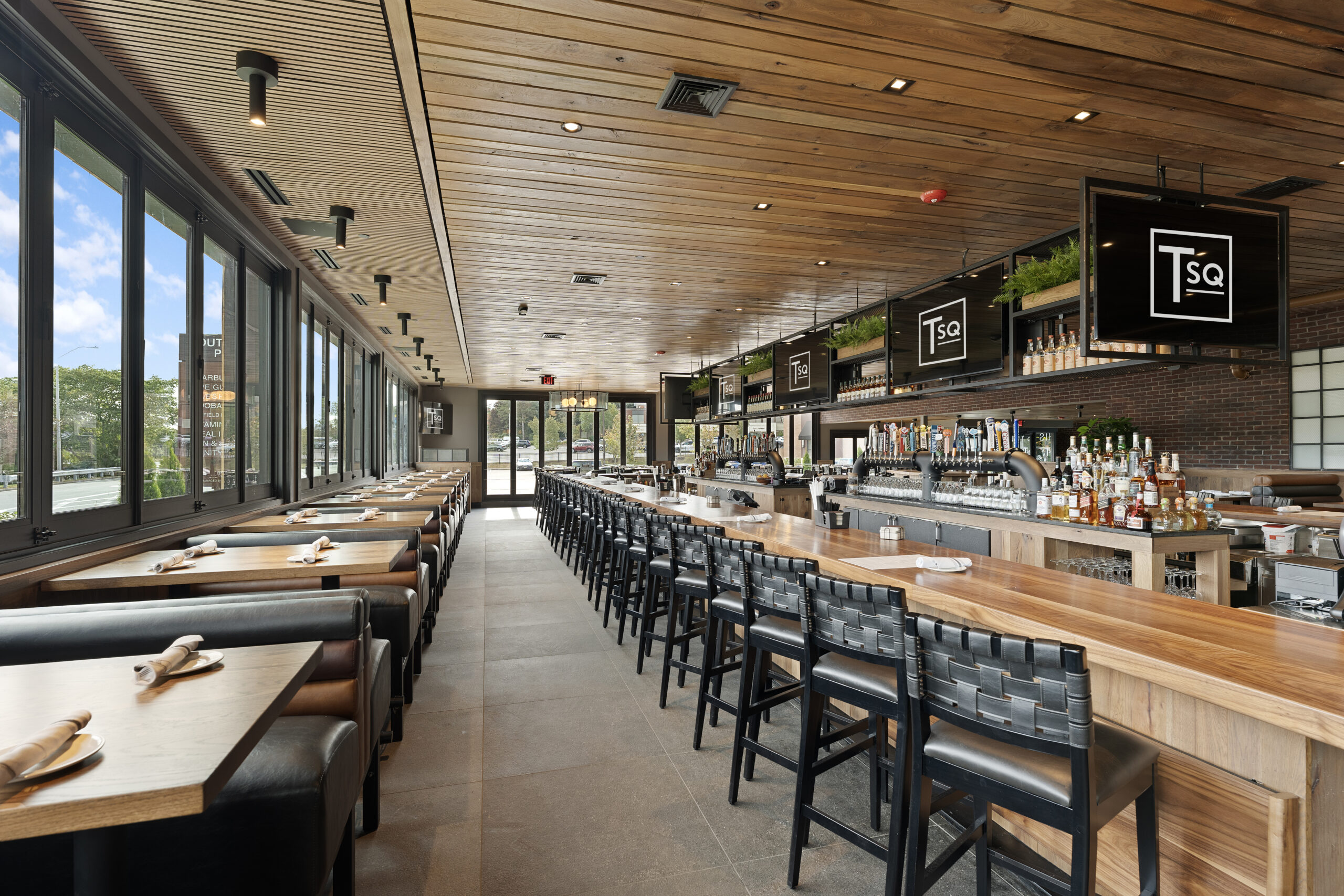 Modern Bar & Restaurant interior with wooden tables, black chairs, a long bar, and large windows providing natural light. TV screens above the bar area display the American Tavern’s logo.