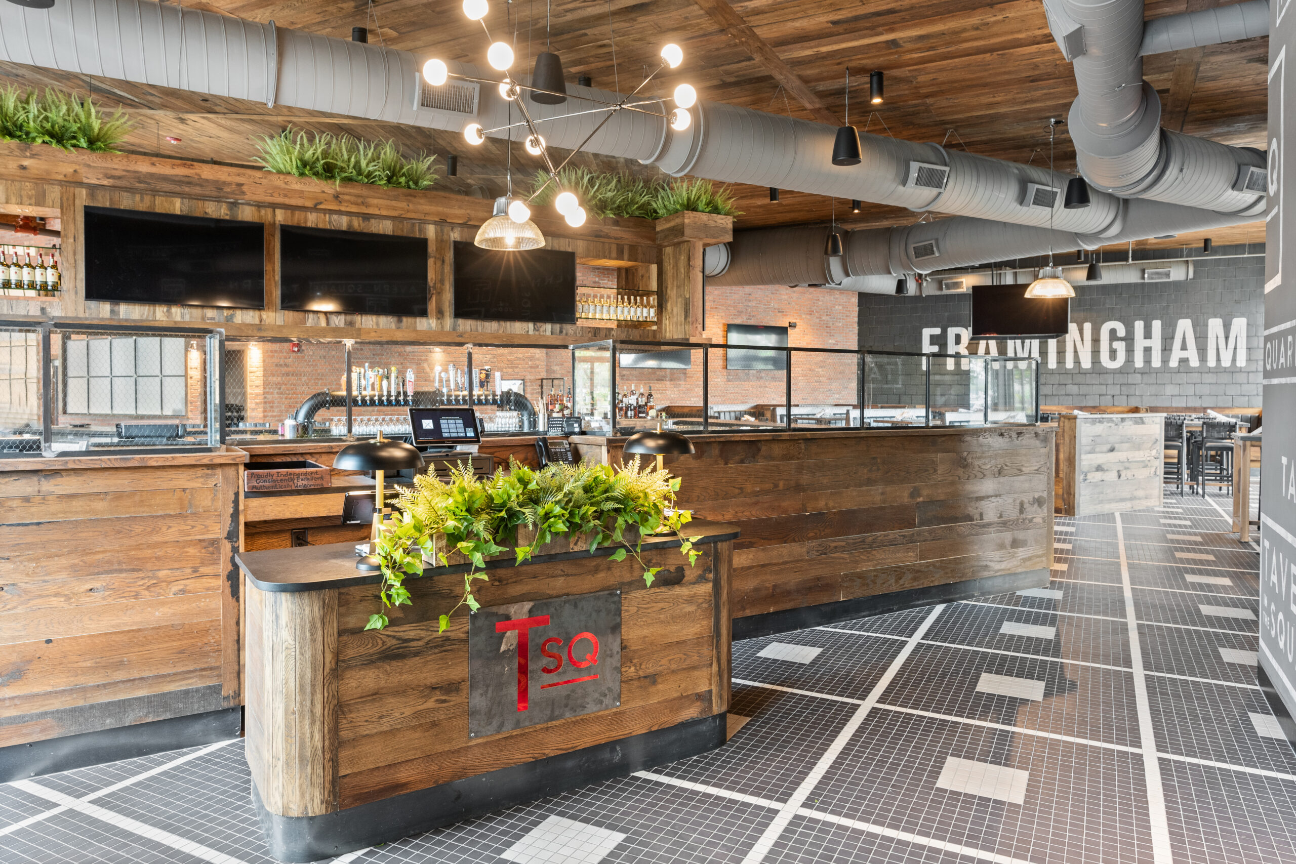 Modern American Tavern interior with wooden décor, a central bar & restaurant counter adorned with plants, large ceiling ductwork, and 