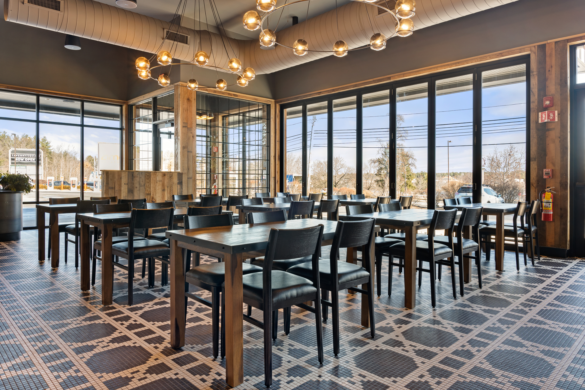 Modern American Tavern interior with wooden tables and black chairs, large windows, tile flooring, and contemporary ceiling lights, with natural light streaming in.