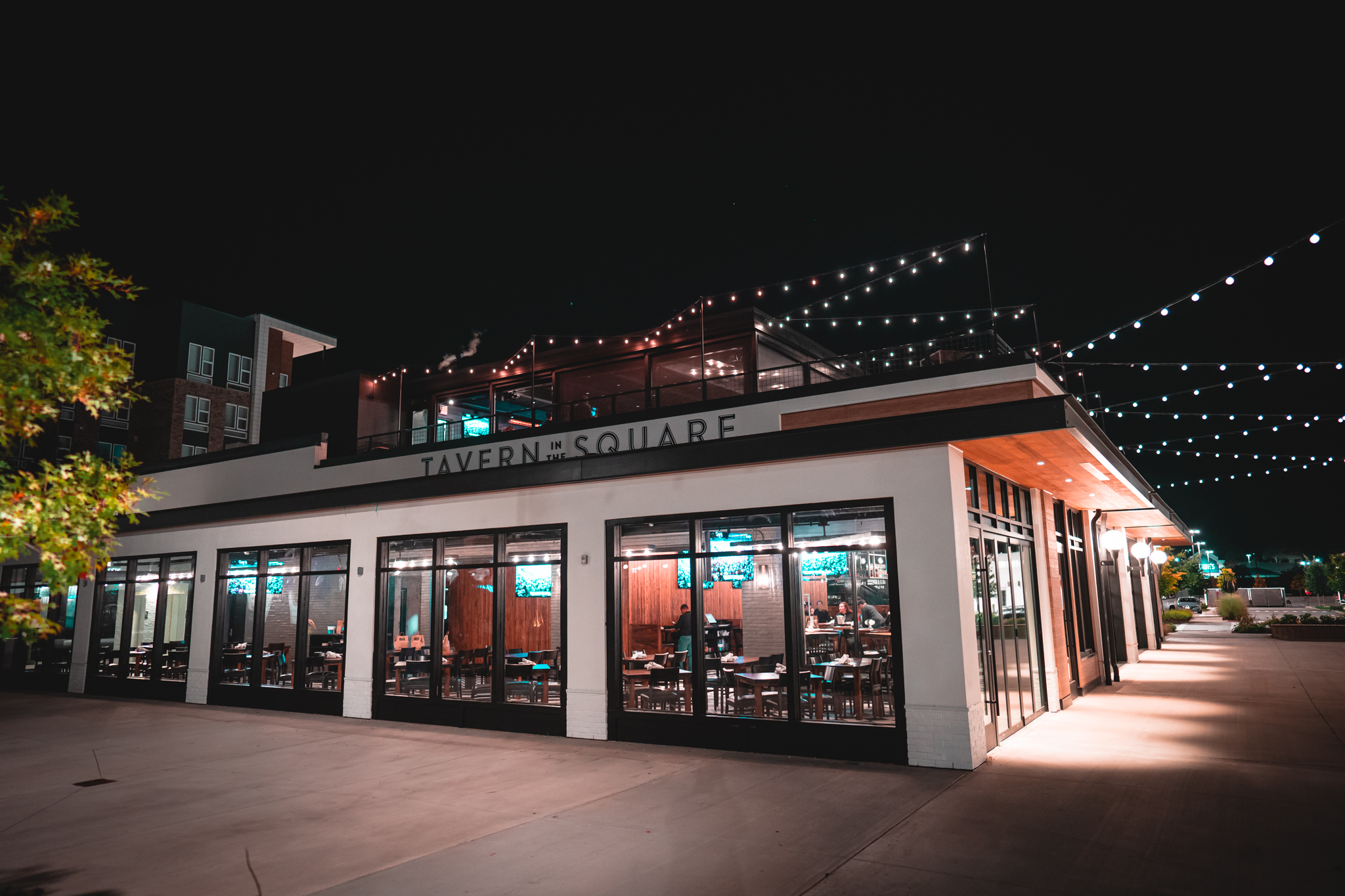 Exterior view of a modern American Tavern called Tavern in the Square at night, with string lights overhead and an empty outdoor seating area, highlighting its inviting Bar & Restaurant vibe.