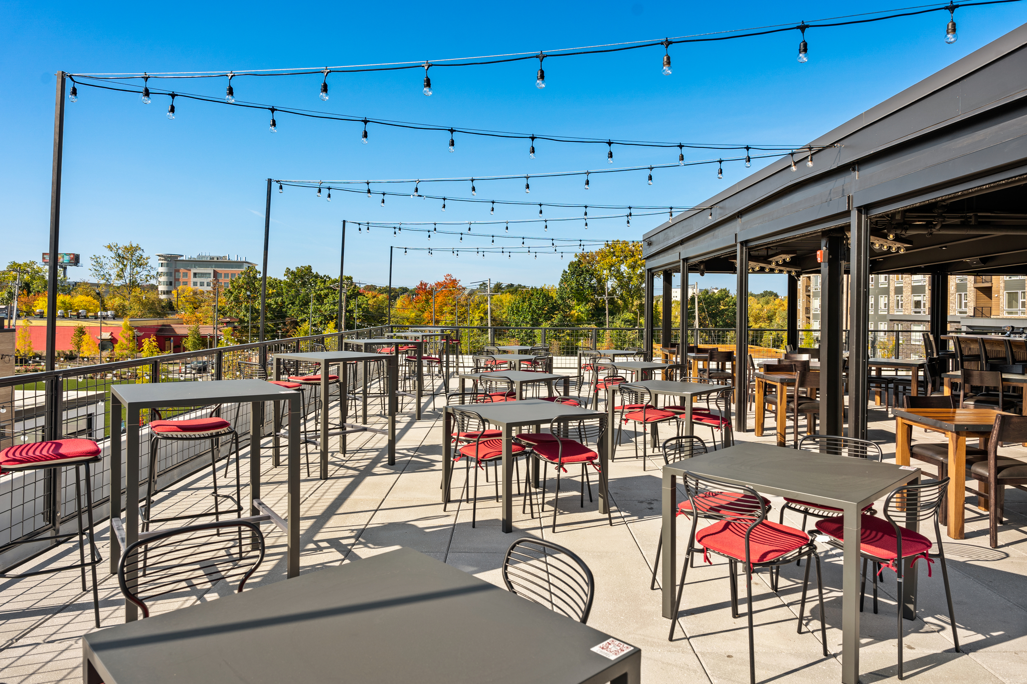 Rooftop patio at this American Tavern features metal tables and red chairs, with string lights overhead and views of trees and buildings—perfect for a sunny day at your favorite Bar & Restaurant.