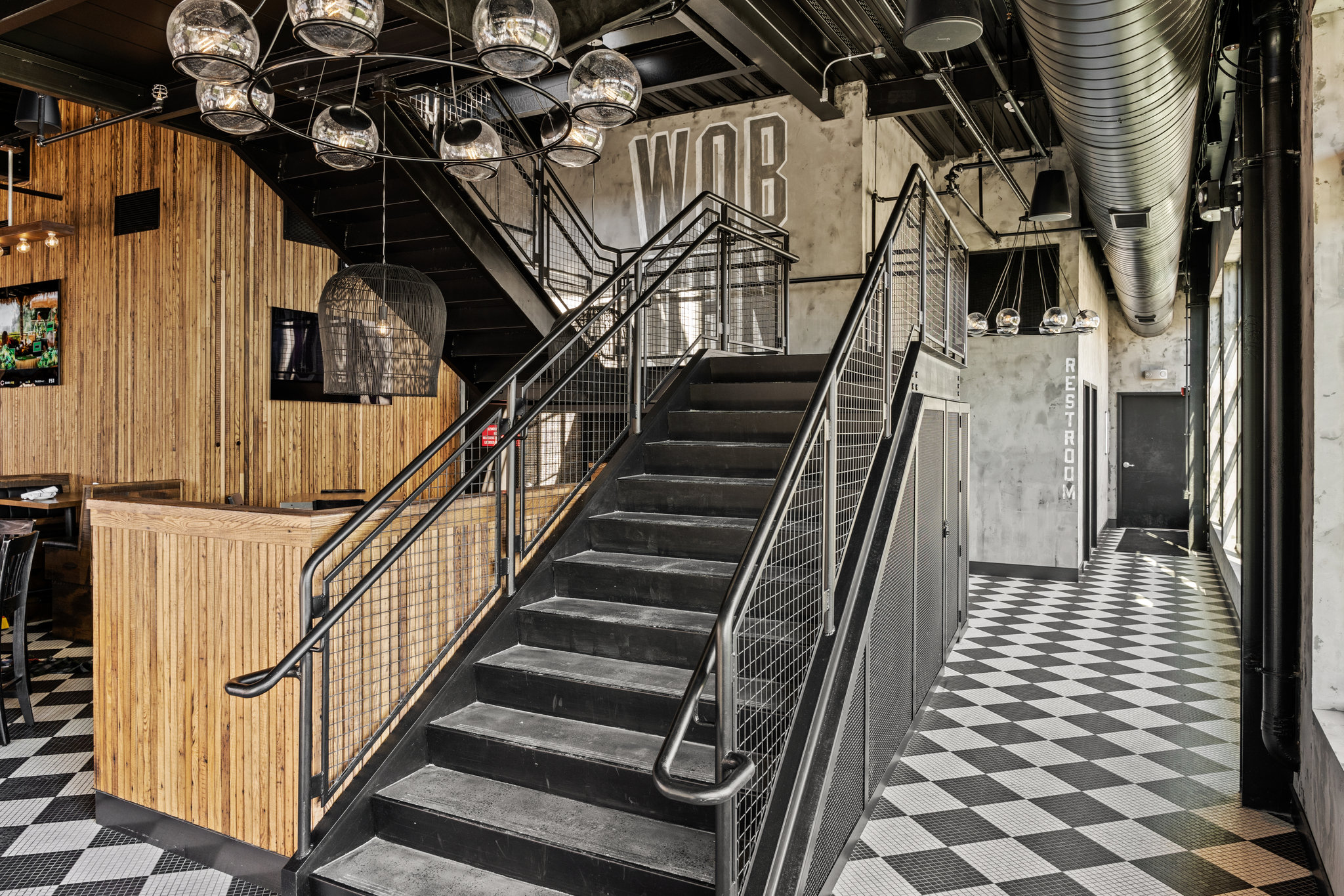 Modern industrial-style interior with black metal staircase, globe pendant lights, checkered tile floor, wood paneling, and signage for the restroom and 