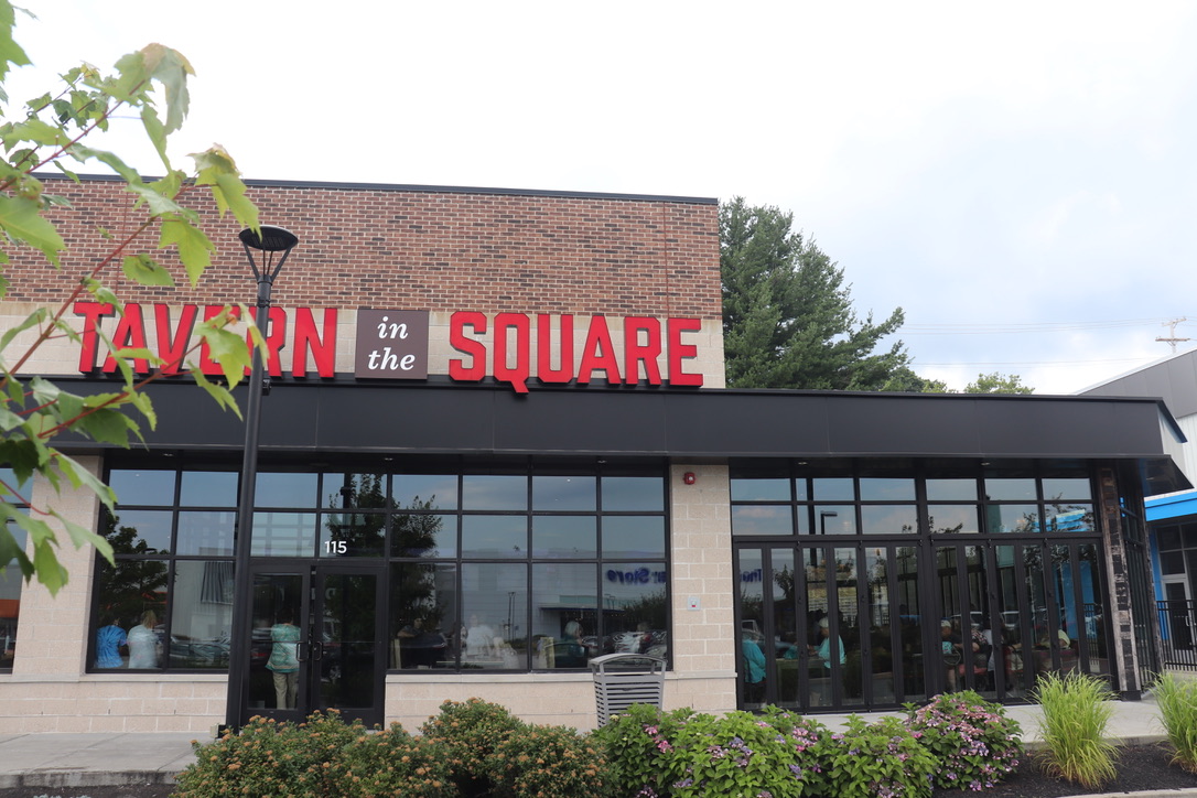 Exterior view of an American Tavern in the Square Bar & Restaurant, featuring large windows, a brick and stone facade, stylish landscaping, and a tree partially in the foreground.