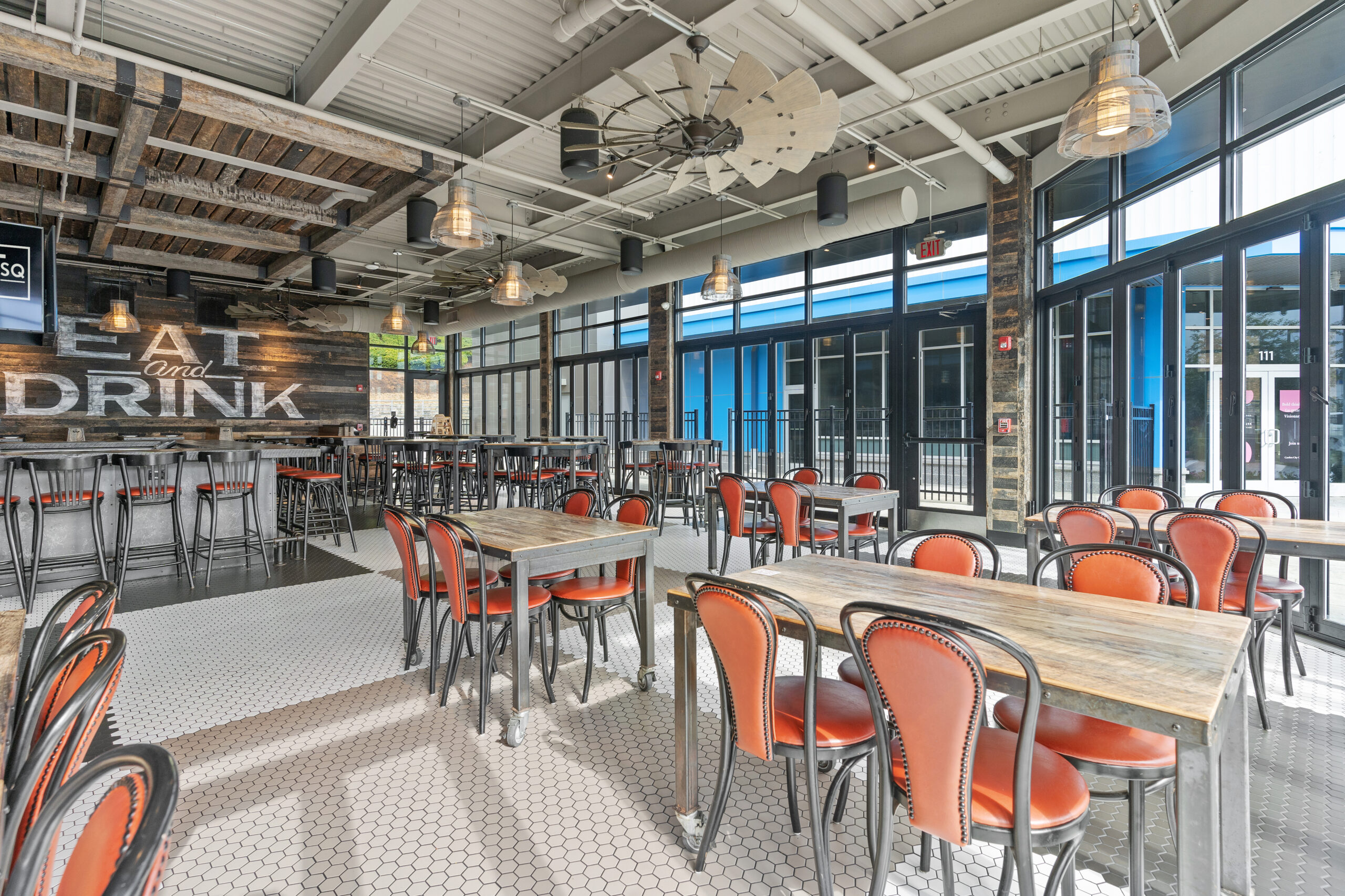 Spacious American Tavern interior with wooden tables, orange-cushioned chairs, tiled floor, and large windows filling the Bar & Restaurant with natural light.