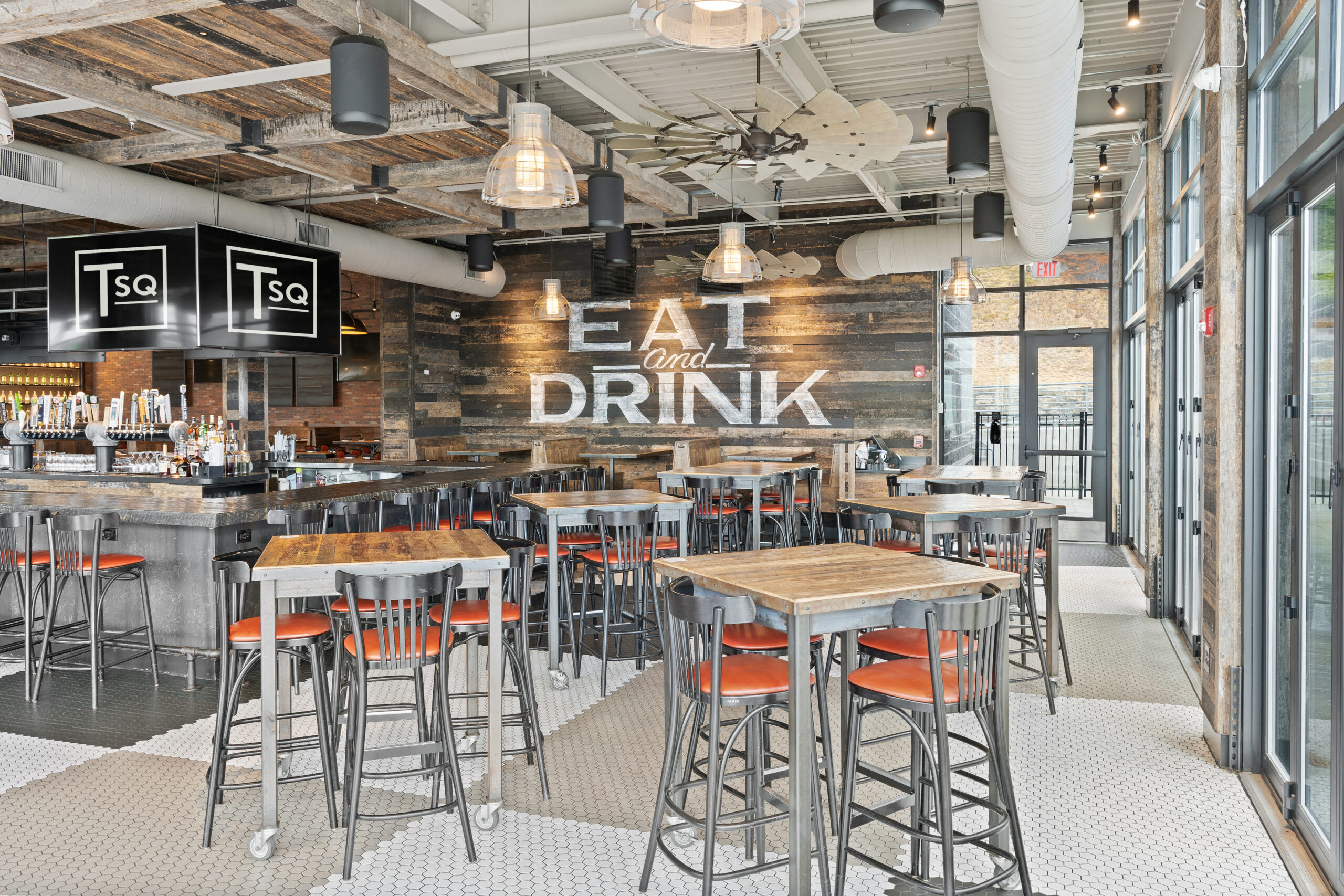 Modern Bar & Restaurant interior featuring wooden tables, metal stools with orange seats, industrial lighting, and a wall sign that reads 
