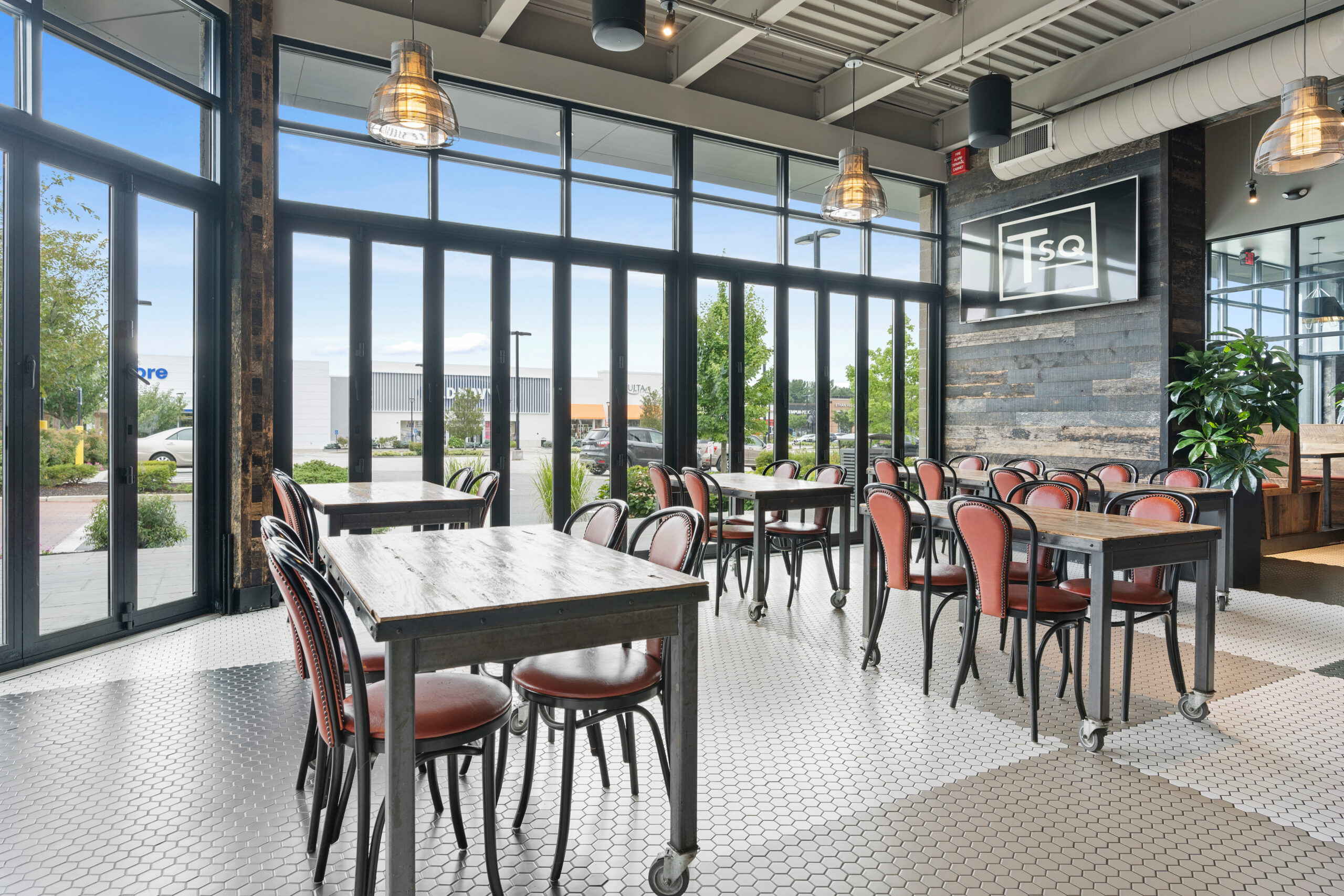 Modern Bar & Restaurant interior with large windows, wooden tables, brown chairs, pendant lights, and white hexagonal tile flooring. Natural light fills the open, spacious dining area for a classic American Tavern feel.