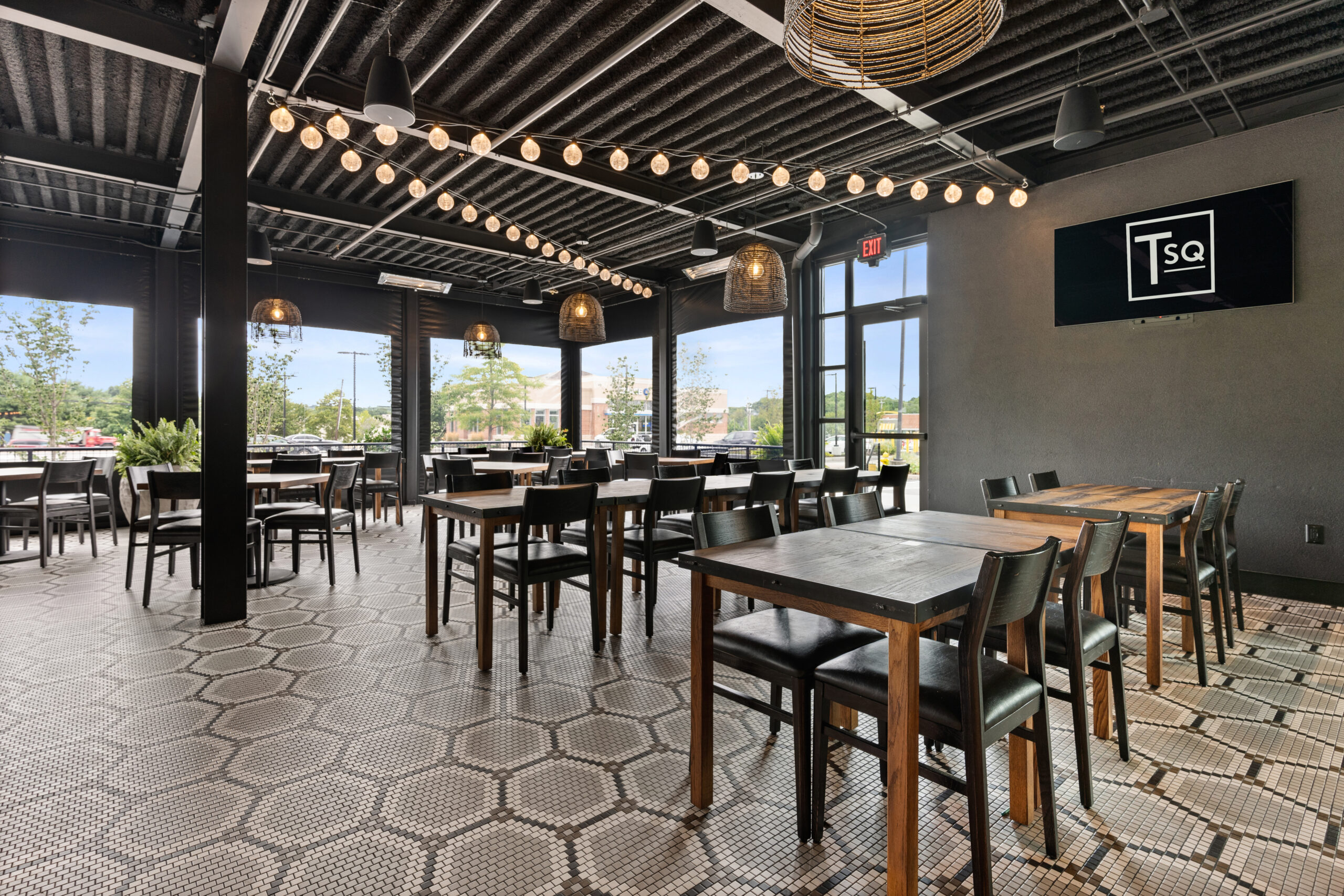 Modern Bar & Restaurant interior with black tables and chairs, string lights, hexagonal tile floor, large windows, and a wall-mounted TV—perfect for an inviting American Tavern experience.
