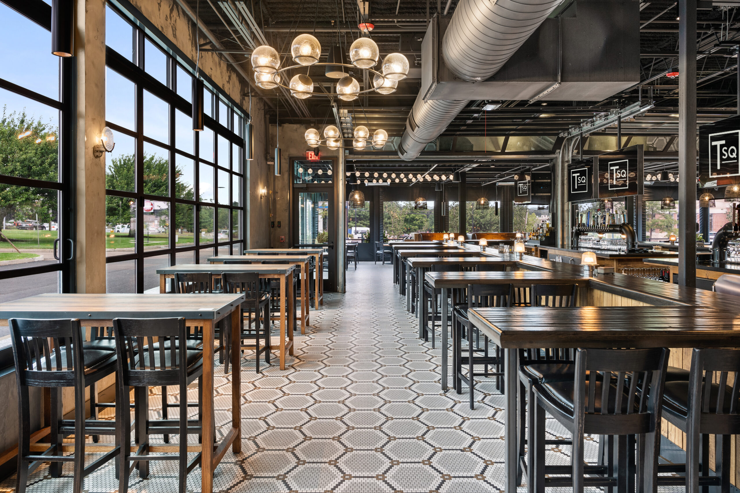Modern American Tavern interior with high tables and chairs, hexagonal tile flooring, large windows, exposed ceilings, and hanging lights—a stylish bar & restaurant atmosphere.