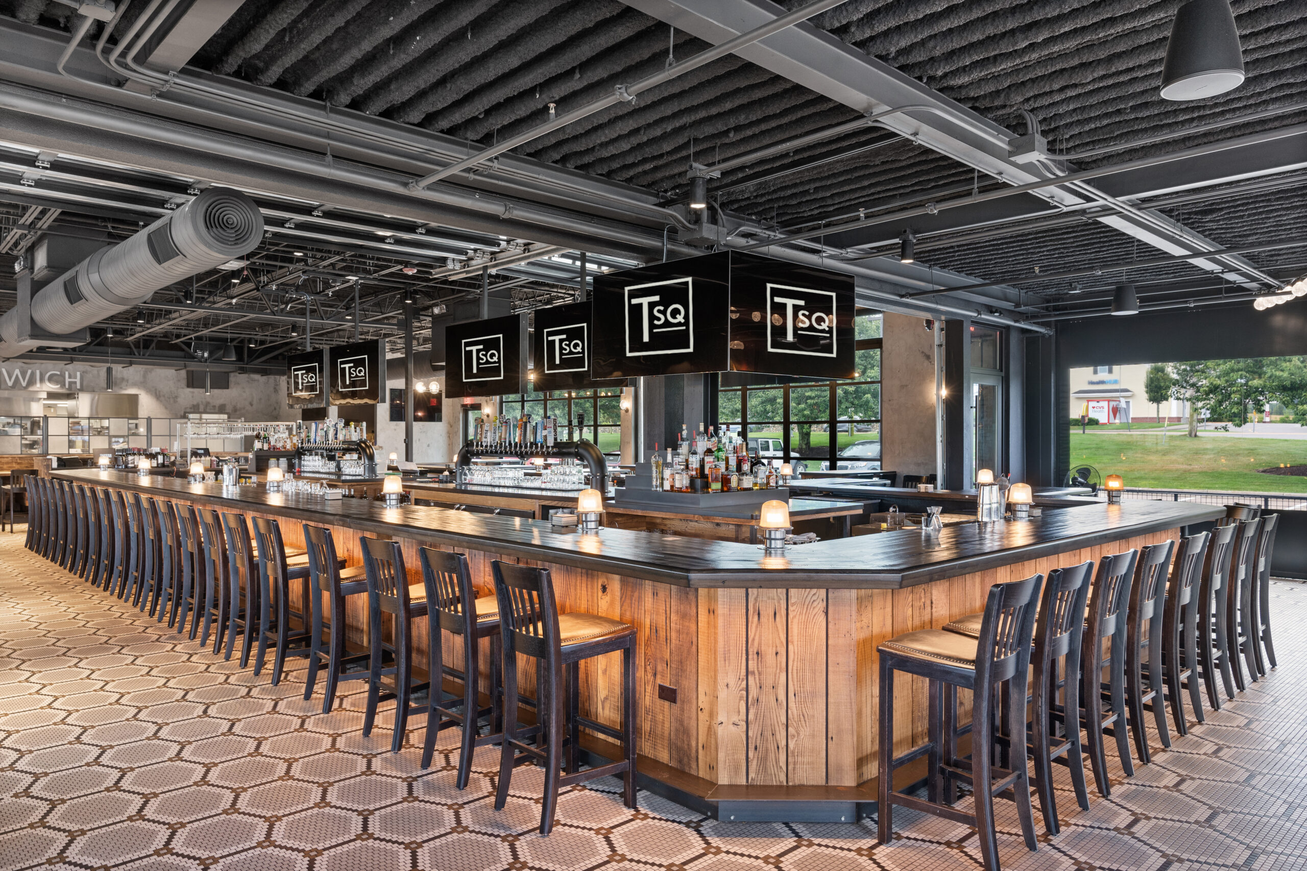 Large modern Bar & Restaurant with a wooden counter, high stools, overhead Tsq signs, and geometric tile flooring; windows reveal lush greenery outside.