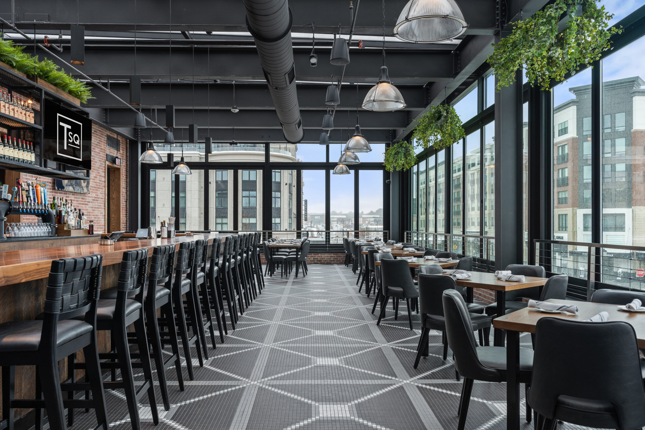 Modern American Tavern interior with large windows, black chairs and tables, a long bar & restaurant area on the left, geometric tile flooring, and hanging plants near the ceiling.