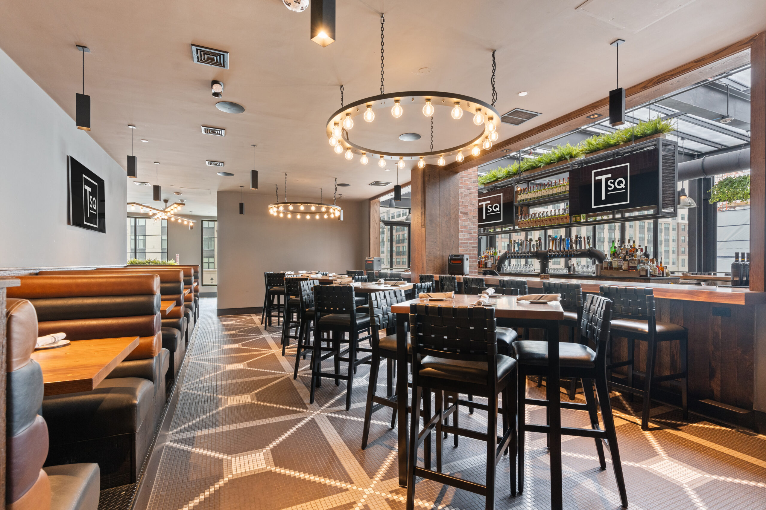 Modern American Tavern interior with geometric tile floor, booth seating on the left, high tables and chairs, circular light fixtures, and a bar area with bottles and greenery overhead.