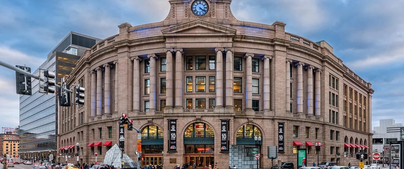 A historic stone train station with columns, a large clock above the entrance, arched windows, and red awnings—now home to a lively American Tavern & Bar & Restaurant—surrounded by cars and pedestrians at a busy intersection.