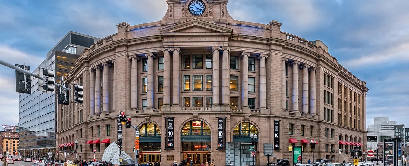 A historic stone train station with columns, a large clock above the entrance, arched windows, and red awnings—now home to a lively American Tavern & Bar & Restaurant—surrounded by cars and pedestrians at a busy intersection.