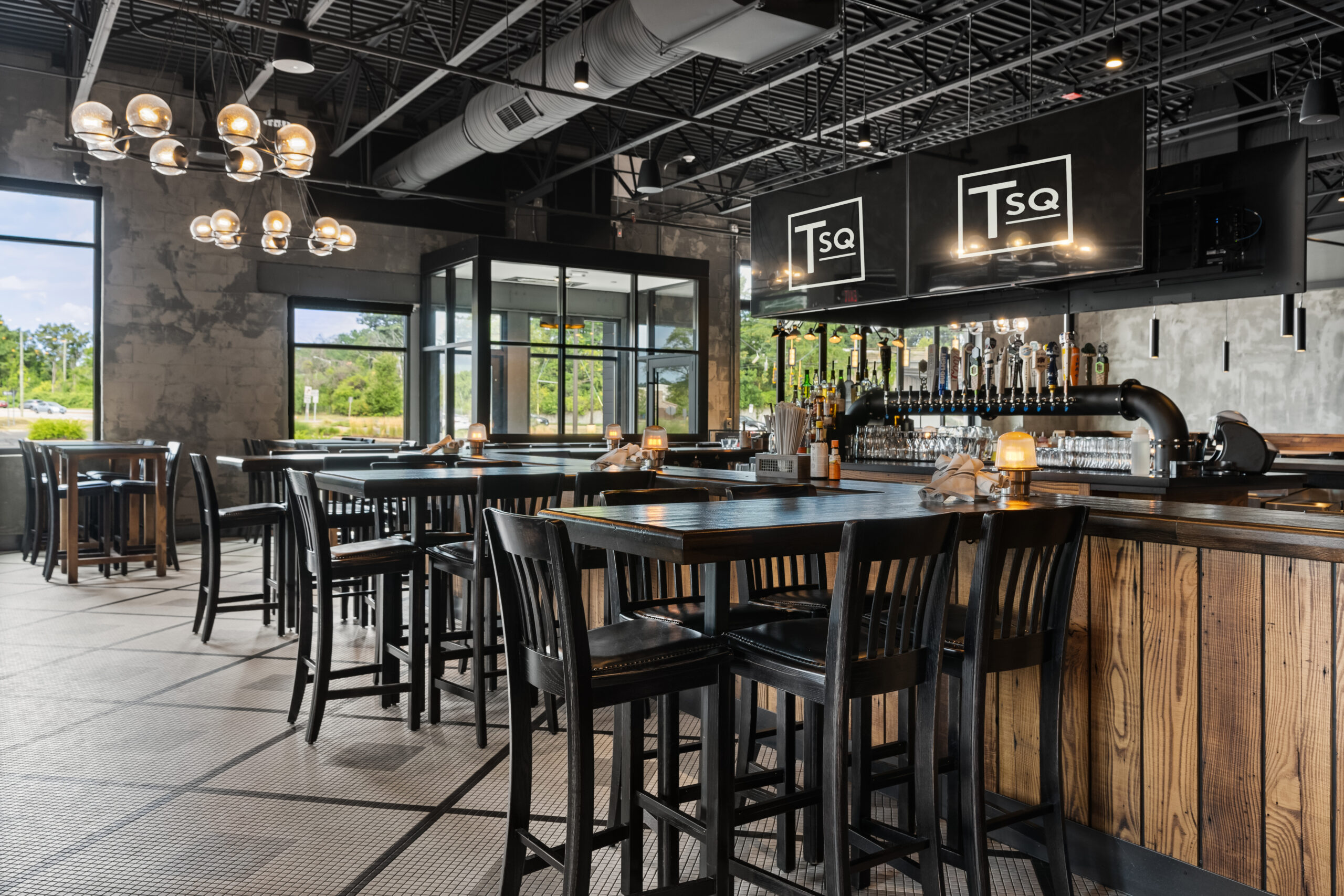 Contemporary Bar & Restaurant interior featuring wooden tables and chairs, hanging light fixtures, large windows, and a bar area with multiple taps and prominent 