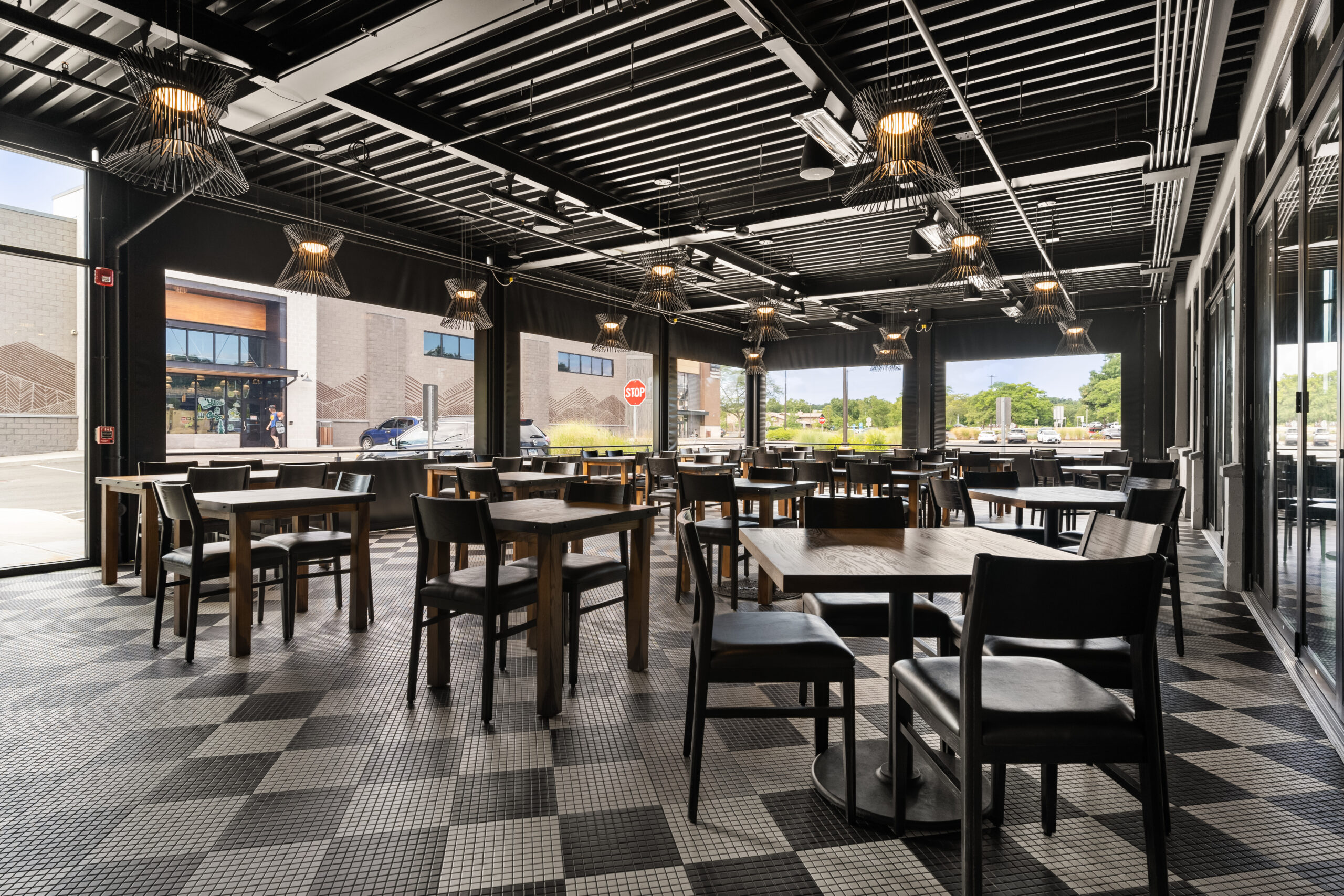 Modern American Tavern interior featuring empty wooden tables, black chairs, large windows, and checkered tile flooring. Daylight streams into this inviting Bar & Restaurant space.