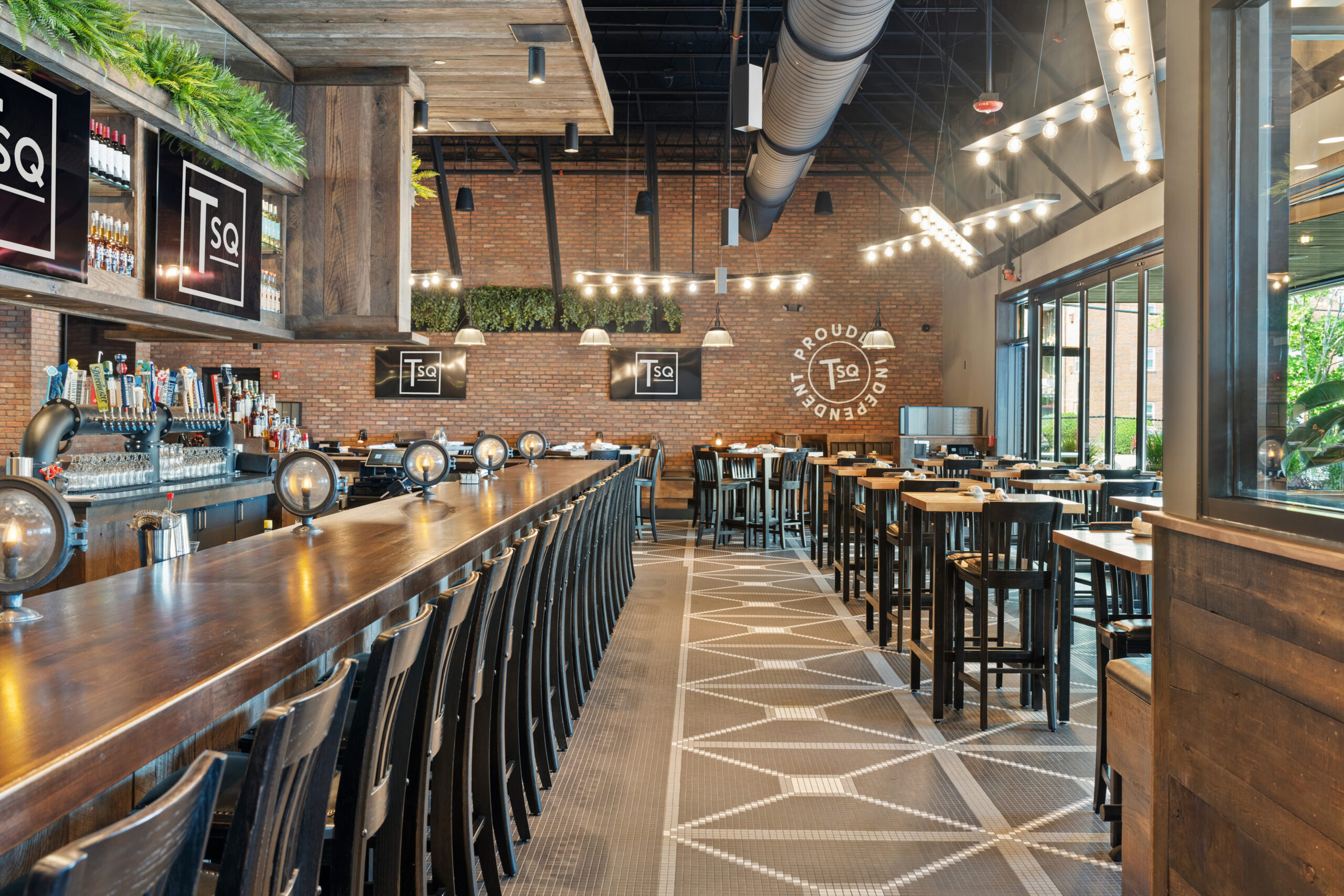 Modern American Tavern bar and restaurant with wooden tables, high chairs, a long bar counter with taps, brick walls, large windows, and multiple TV screens displaying the “TSq” logo.