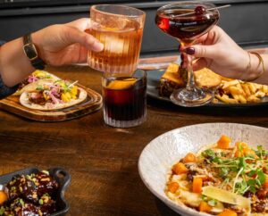 Two people clink cocktail glasses over a wooden table set with tacos, fries, a sandwich, a salad, and glazed appetizers—perfect Winter Pairings for a cozy gathering.