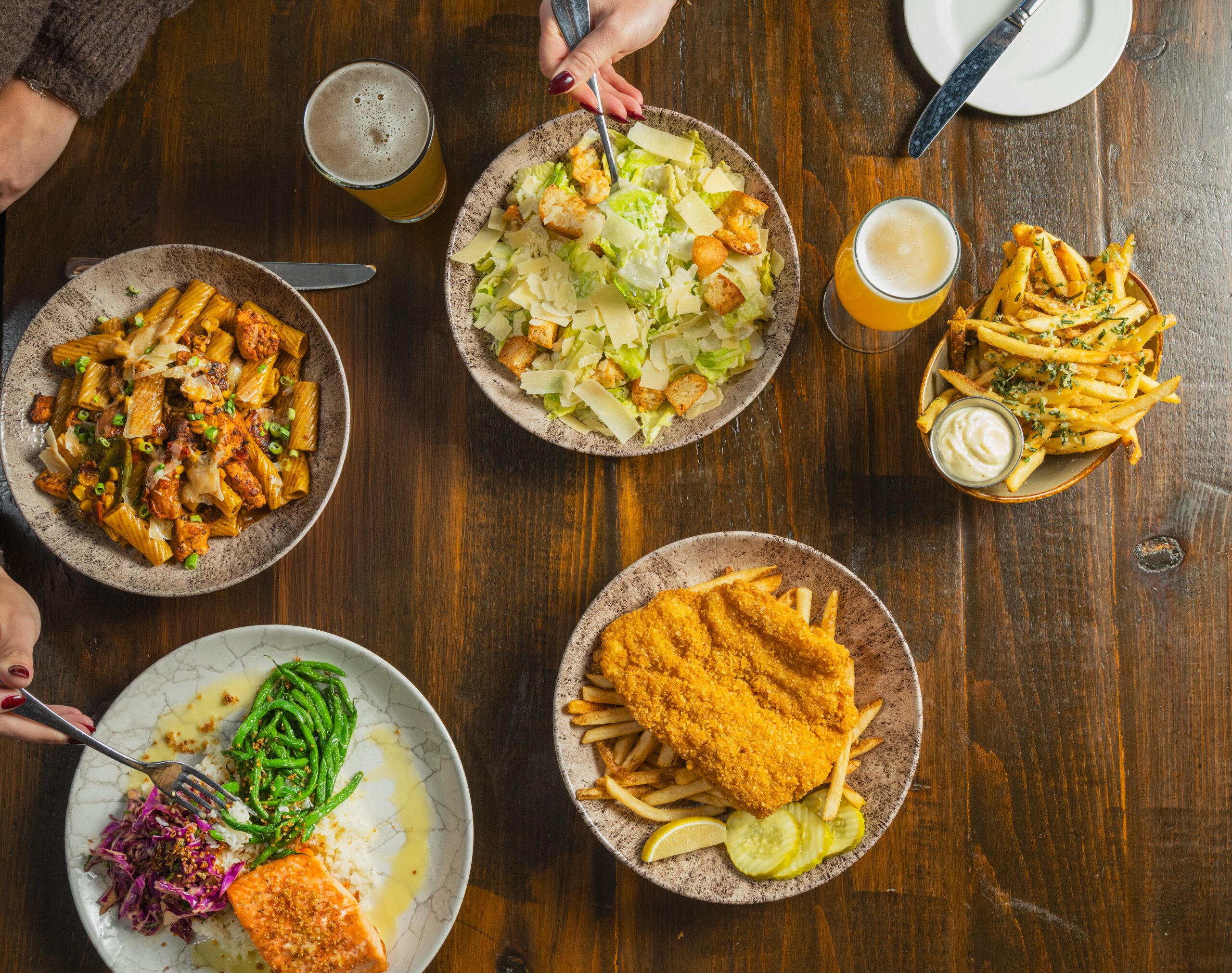 A wooden table set with plates of pasta, Caesar salad, fries, fried fish with sides, and two glasses of beer at an inviting American Tavern Bar & Restaurant, with hands reaching for food.