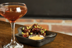 A cocktail in a stemmed glass sits next to a black dish of glazed chicken wings garnished with green onions and a small cup of dipping sauce—perfect Winter Pairings for cozy evenings.