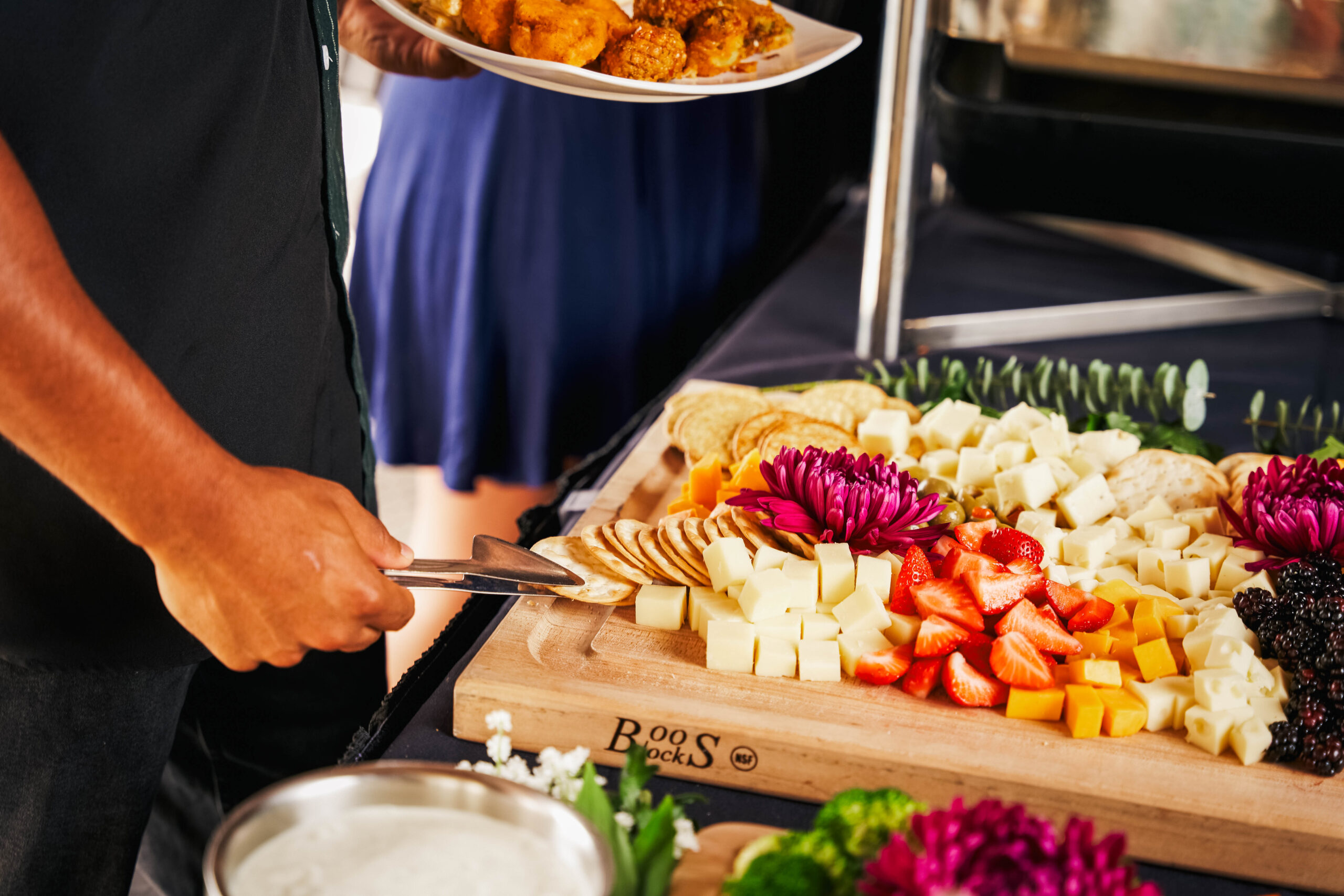A person uses tongs to select cheese and crackers from a wooden platter with assorted cheeses, crackers, strawberries, and decorative flowers at an inviting American Tavern Bar & Restaurant.