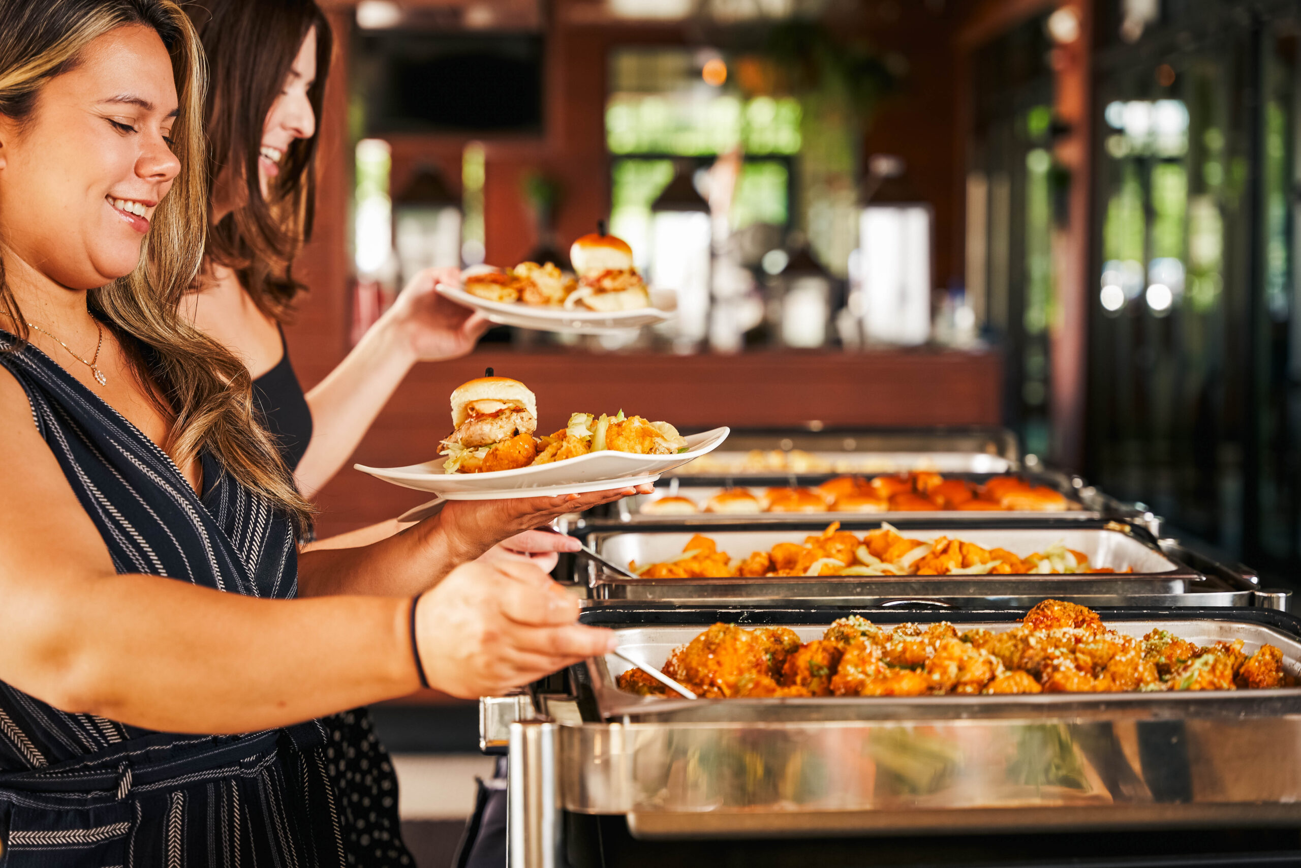 Two women serve themselves food from a buffet with hot trays, selecting sliders and fried chicken in a bright, casual American Tavern-style Bar & Restaurant.