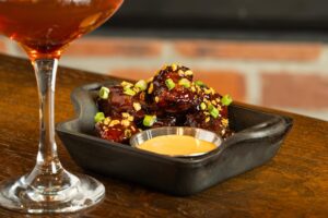 A cocktail in a coupe glass sits next to glazed short rib bites topped with green onions and a side of dipping sauce in a black dish on a wooden table, making for delicious Winter Pairings.