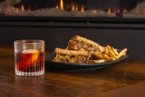 A glass of cocktail sits next to a plate of grilled sandwich filled with short rib meat and cheese, served with French fries, on a wooden table in front of a fireplace—perfect Winter Pairings for a cozy night in.
