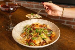 A hand holds a fork with pasta above a bowl topped with short rib, greens and sun-dried tomatoes, next to a cocktail on a wooden table in front of a fireplace—perfect for cozy Winter Pairings.