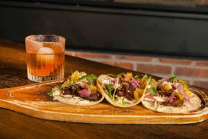A wooden board with three loaded shot rib tacos topped with onions, arugula, and cheese sits next to a glass of cocktail with ice and a salted rim—perfect for cozy Winter Pairings on a dark wooden table.