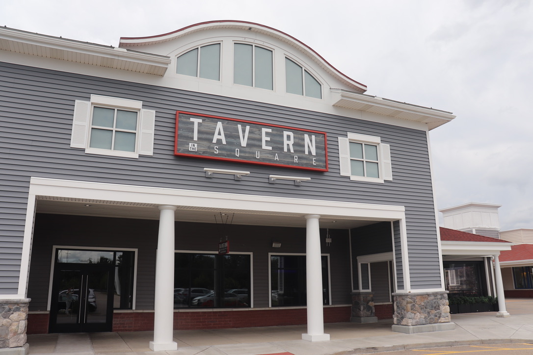 A building with gray siding and stone columns features a large sign reading “TAVERN SQUARE” above the entrance, hinting at a welcoming American Tavern bar & restaurant atmosphere inside.