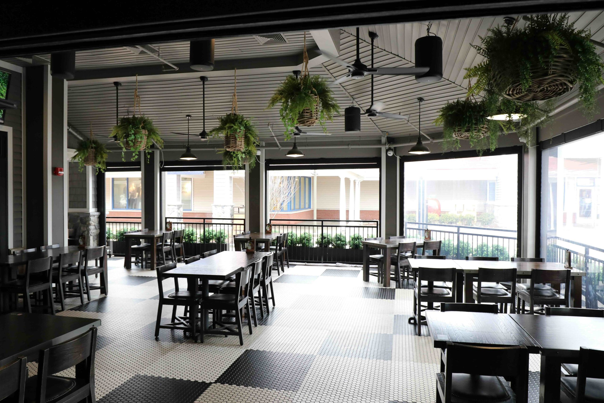 A spacious indoor dining area in Wrentham MA with black tables and chairs, tiled floor, hanging plants, large windows, and views of an outdoor walkway.