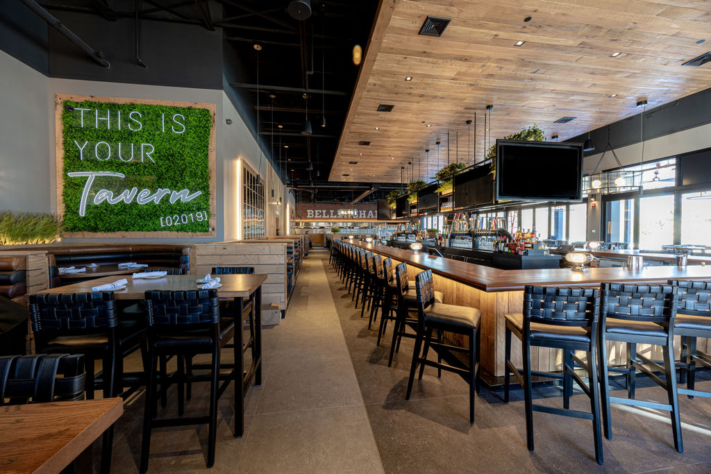 Modern tavern interior with wooden tables, high chairs, and a large bar evoke the welcoming vibe of Tavern in the Square. A green wall sign reads 