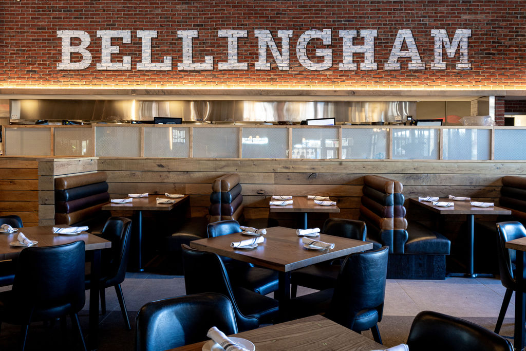 Modern restaurant interior with wooden tables, black chairs, and booth seating; “BELLINGHAM” is displayed in large white letters on a brick wall above the open kitchen area at Tavern in the Square.